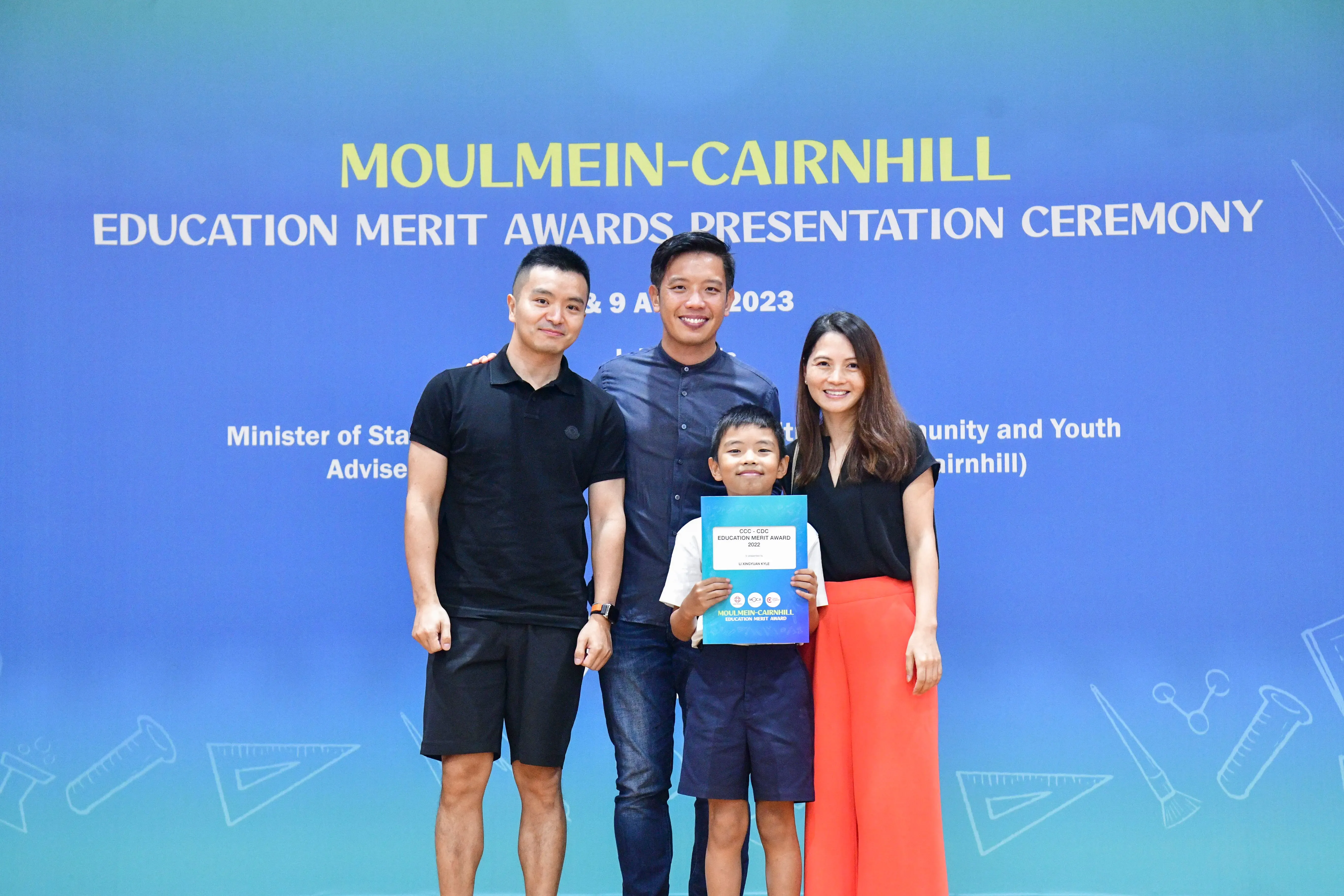 Three adults and a young boy holding an award certificate pose in front of a Moulmein-Cairnhill Education Merit Awards banner.
