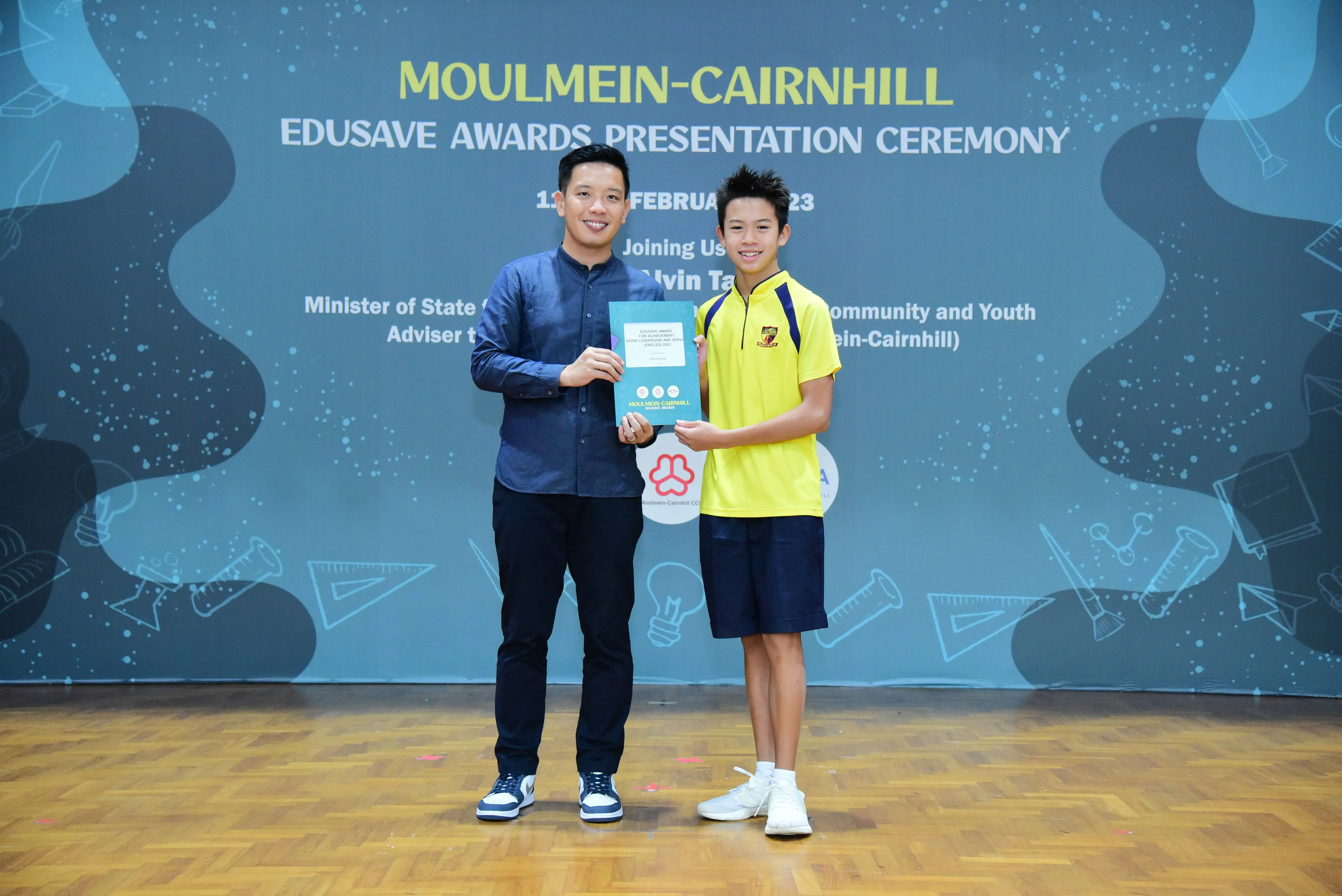 Two people hold a certificate together at the Moulmein-Cairnhill Edusave Awards Presentation Ceremony backdrop.