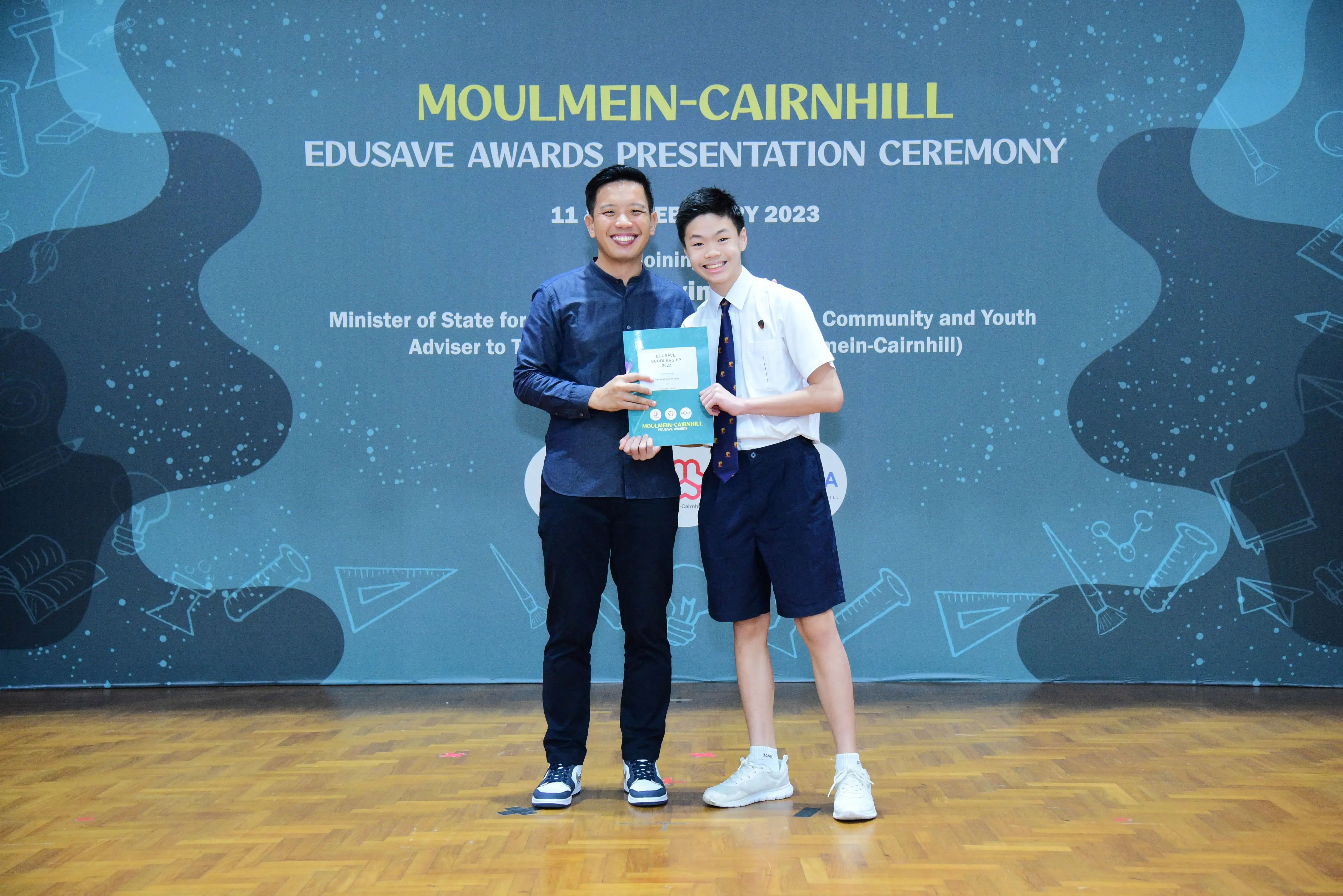 An adult and a student in school uniform hold an award certificate at the Moulmein-Cairnhill Edusave Awards Ceremony 2023.