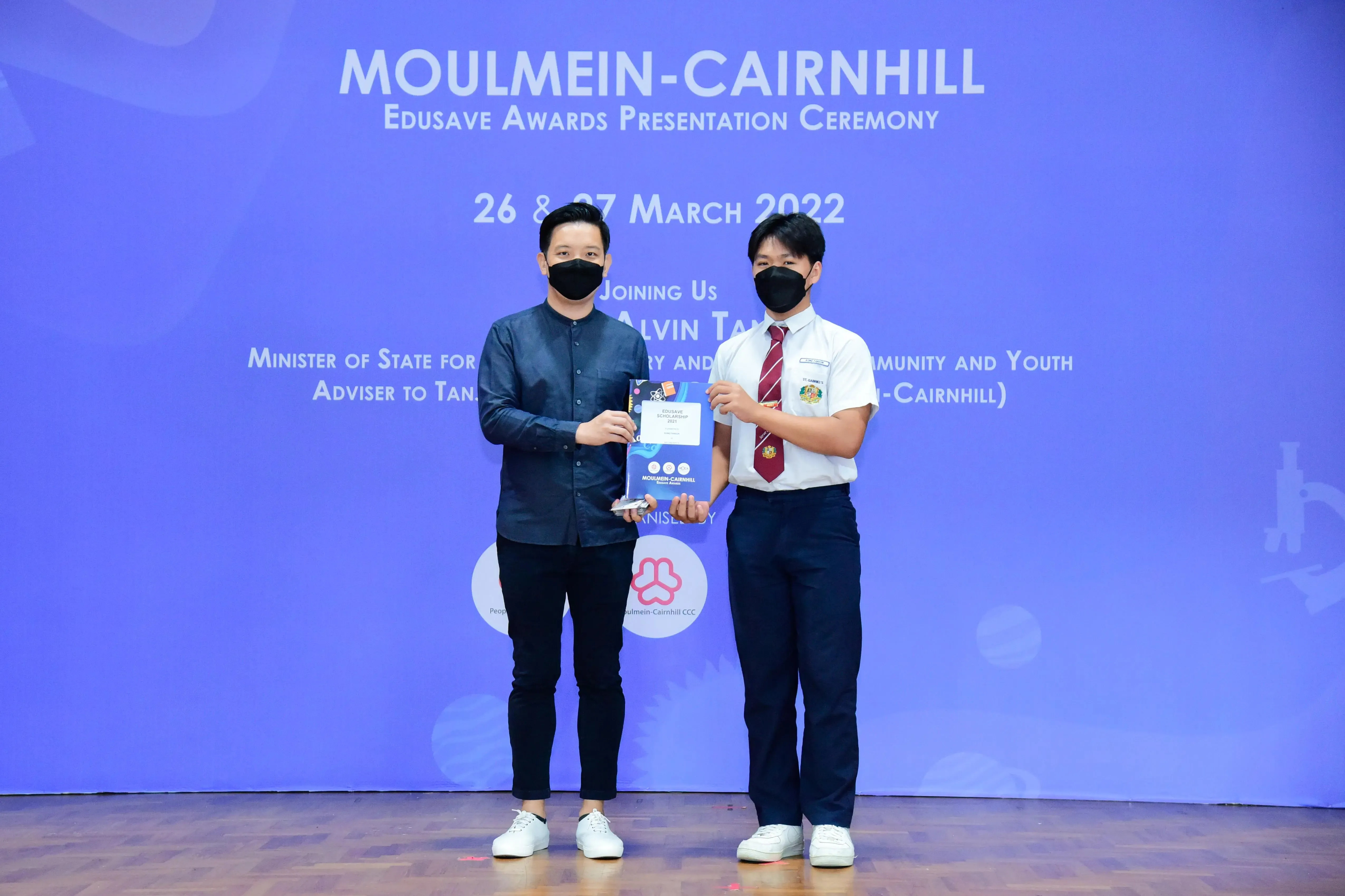 Two masked men hold an award on stage at the Moulmein-Cairnhill Edusave Awards Ceremony, March 2022.
