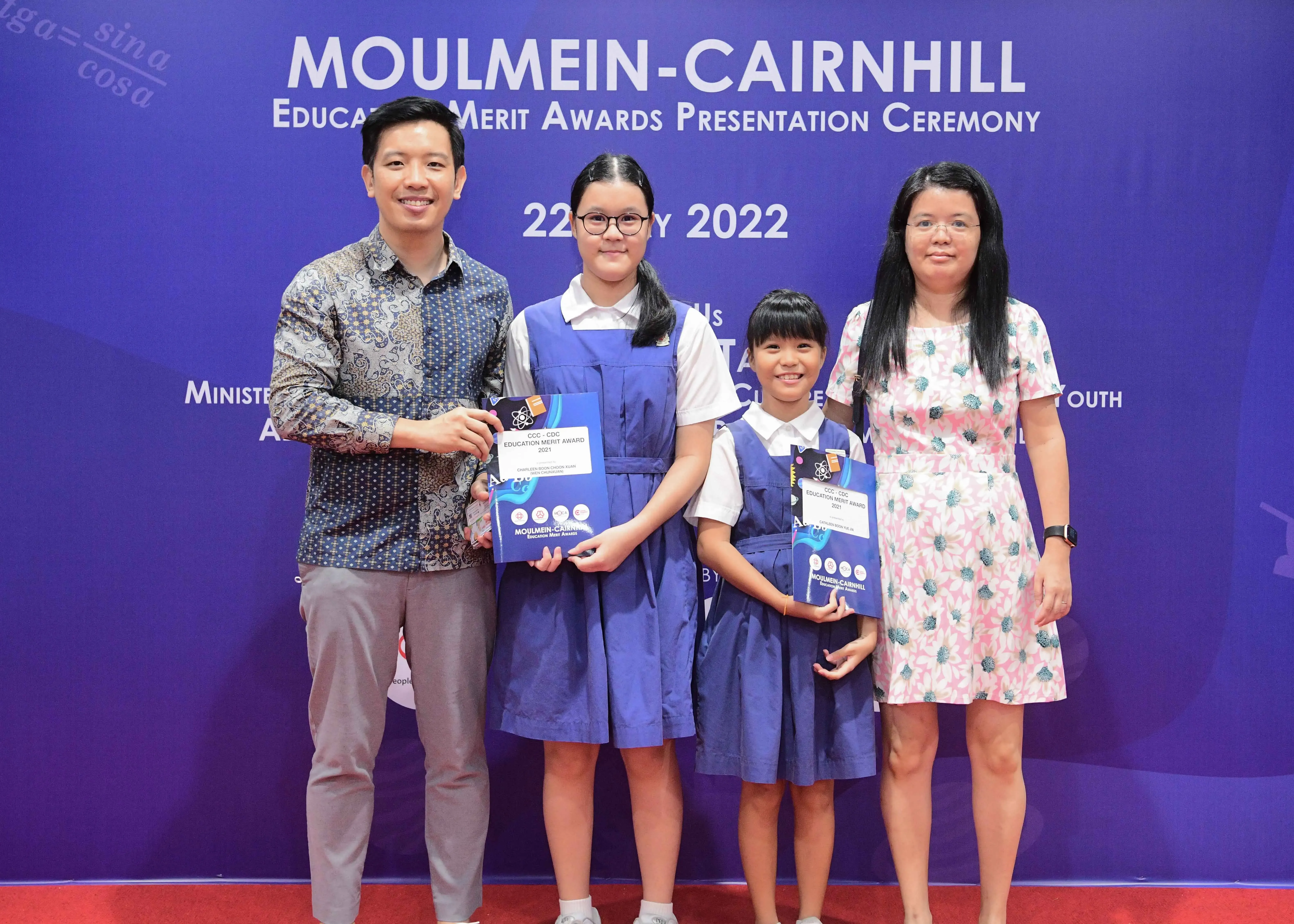 Four people pose before a Moulmein-Cairnhill Education Merit Awards 2022 banner, two girls in school uniforms holding award certificates.