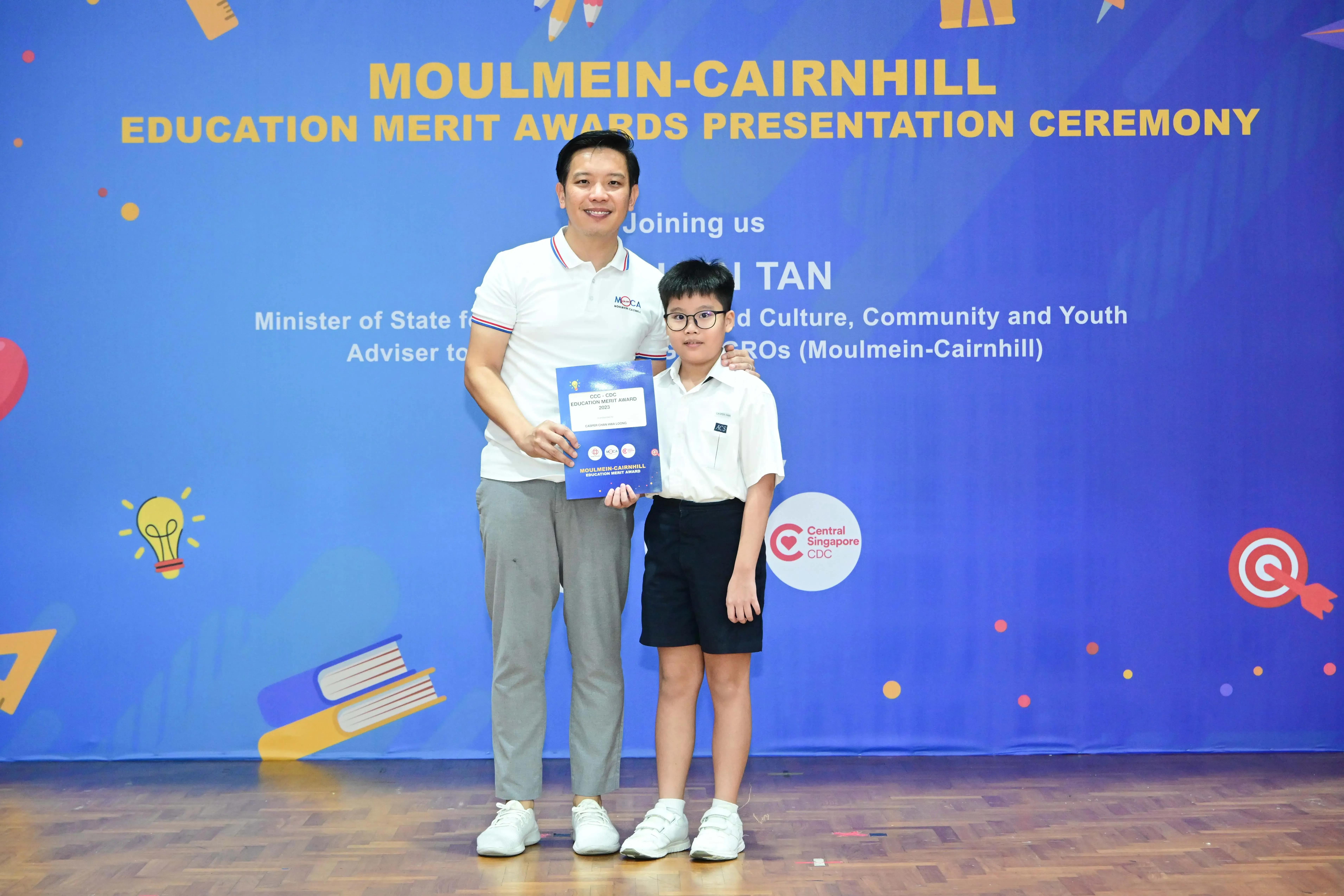 A man and a young boy pose holding an award booklet at the Moulmein-Cairnhill Education Merit Awards ceremony.