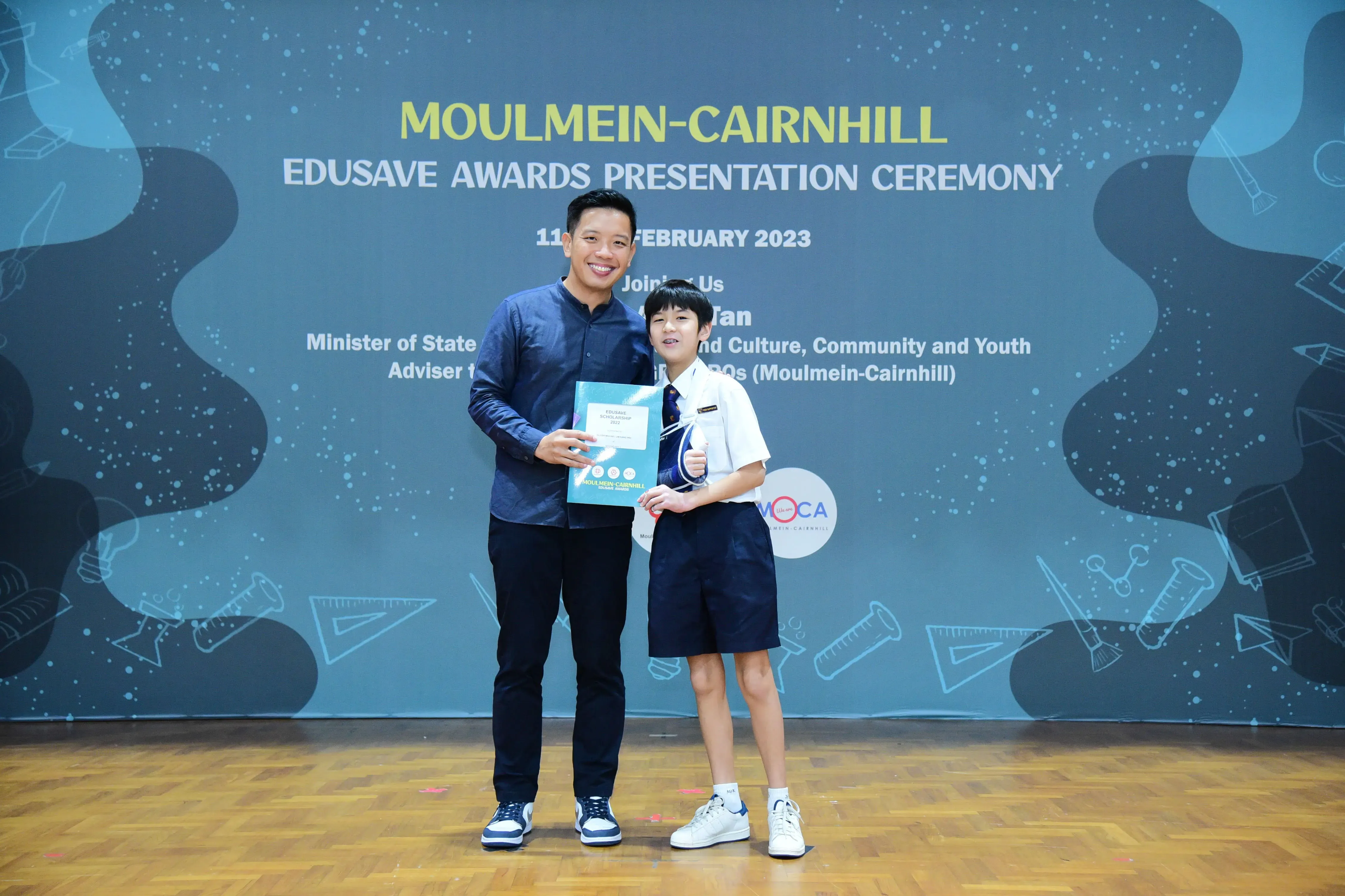 An adult and a student in school uniform hold a certificate at the Moulmein-Cairnhill Edusave Awards Ceremony, February 2023.