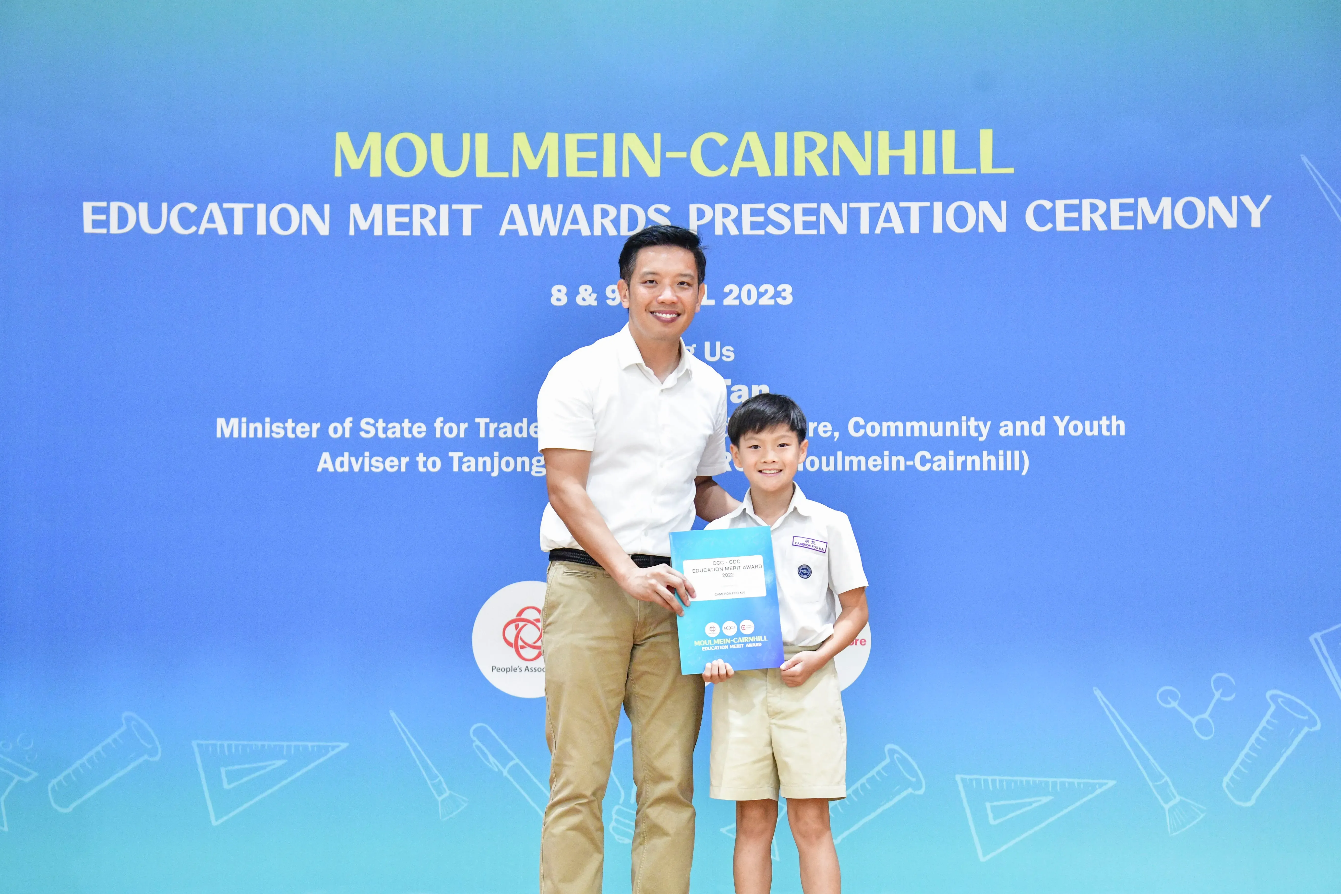 A man and a young boy in school uniform hold an award certificate in front of a blue Moulmein-Cairnhill ceremony backdrop.