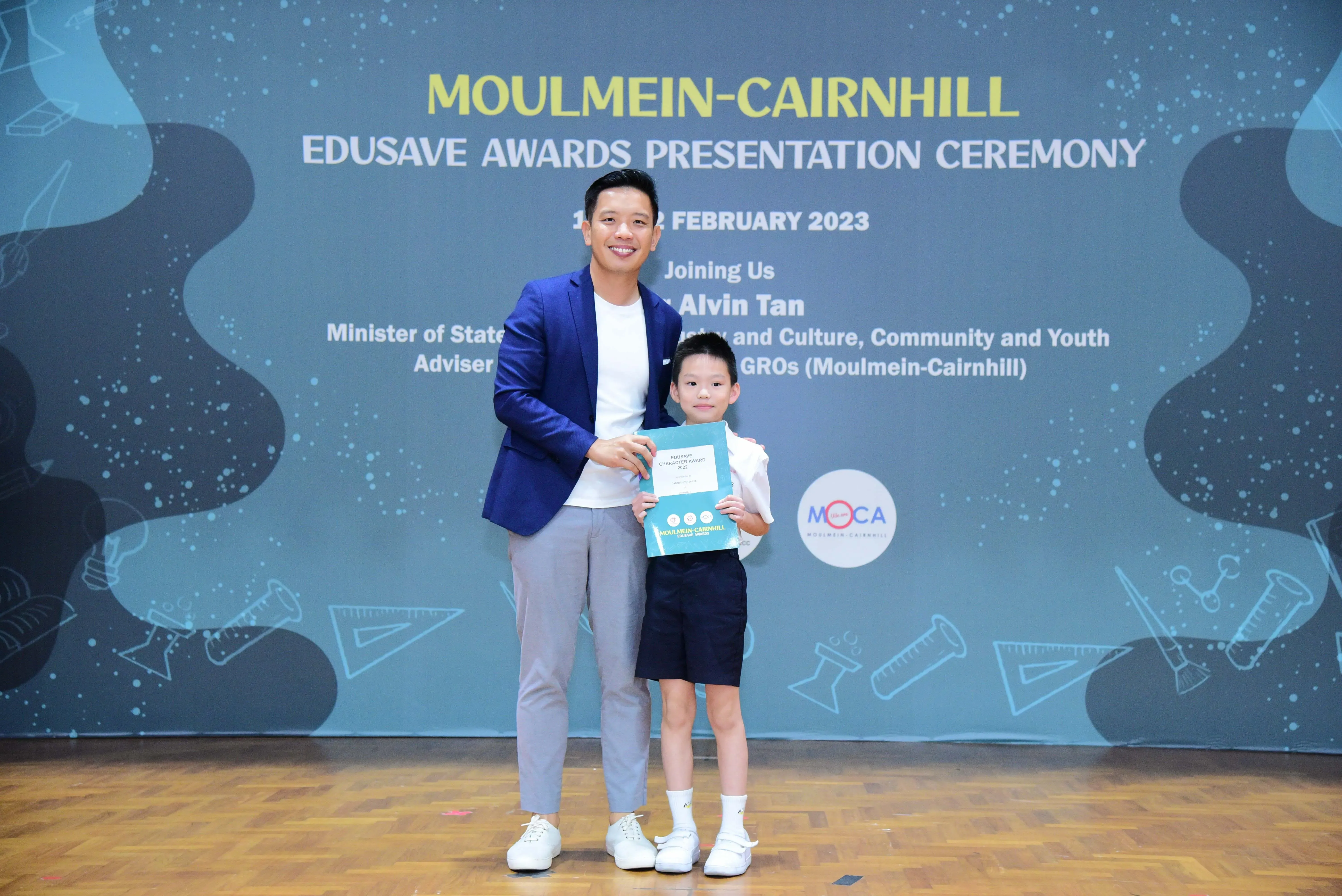 A man in a blue blazer and a young boy hold a certificate at the Moulmein-Cairnhill Edusave Awards Ceremony 2023.