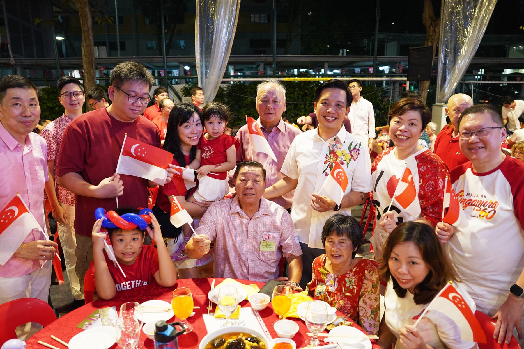 A large group of smiling people in red and white clothing hold Singapore flags at an outdoor dinner celebration.