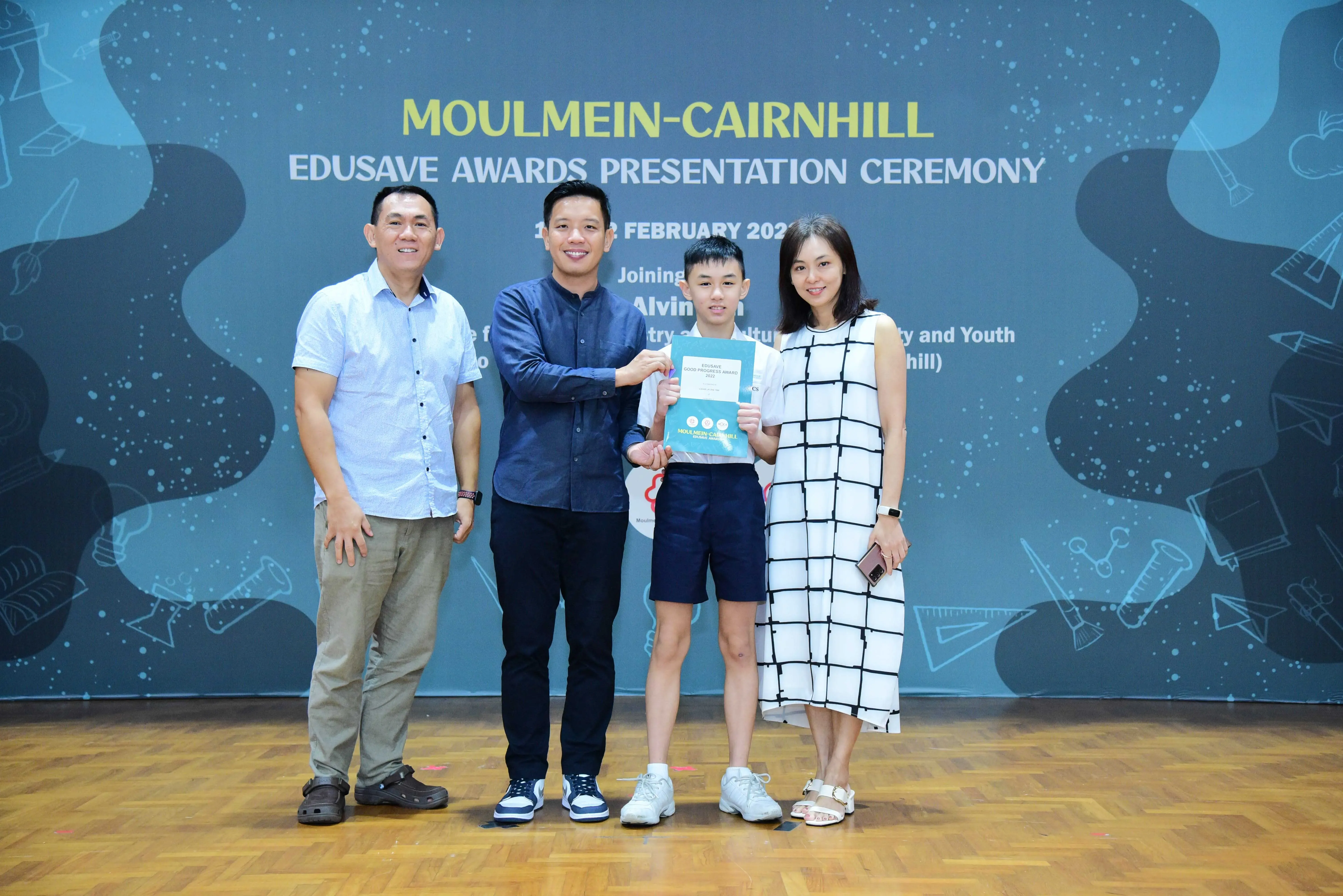 Four people pose with a certificate at the Moulmein-Cairnhill Edusave Awards Presentation Ceremony backdrop.