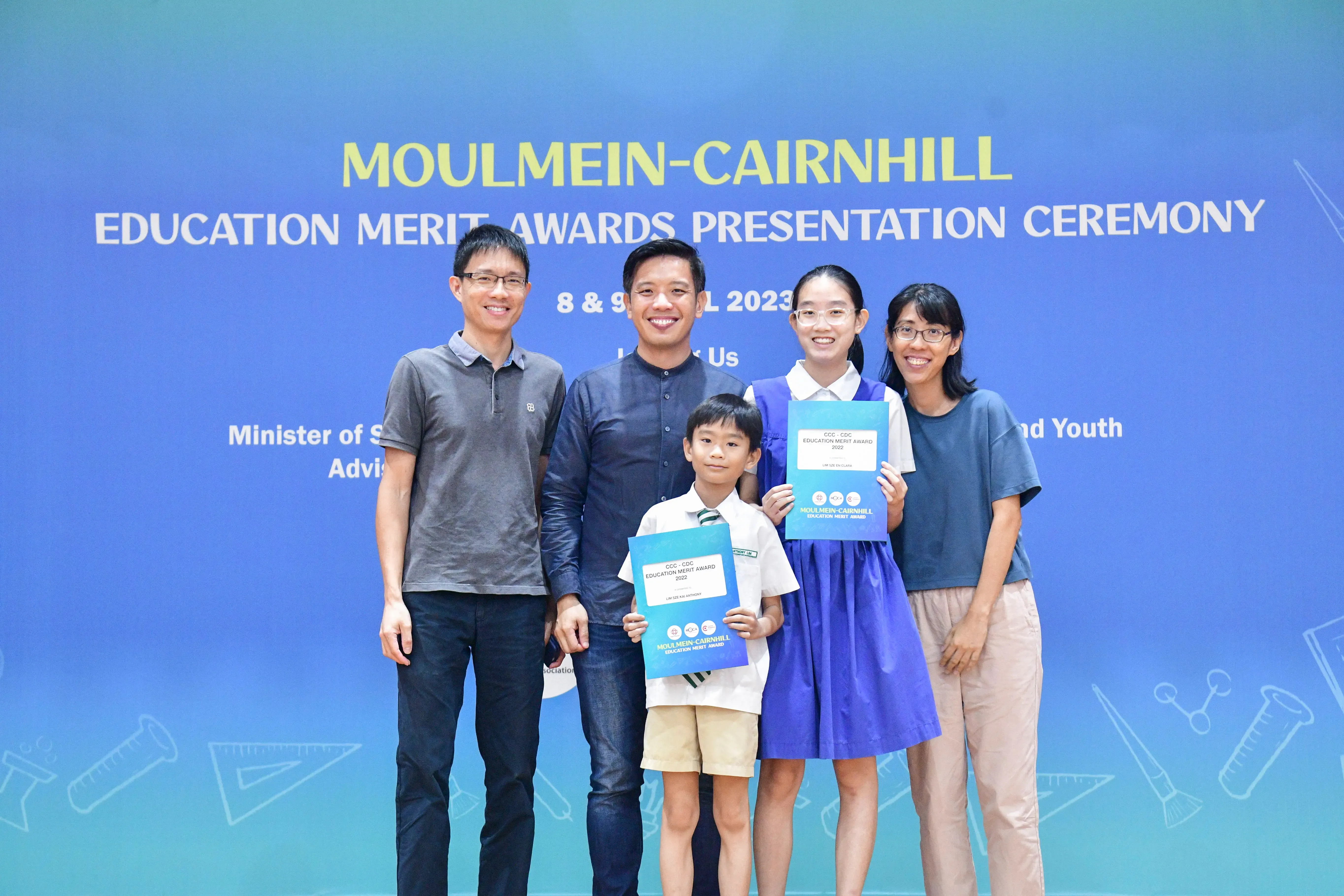 Five people pose in front of a blue Moulmein-Cairnhill Education Merit Awards Presentation Ceremony banner.
