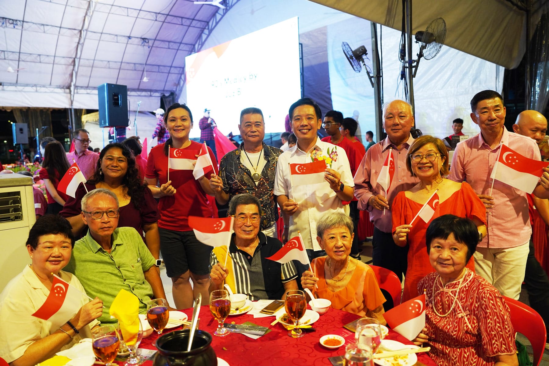 Group of smiling people holding small Singapore flags at an outdoor event with a red tablecloth and drinks nearby.