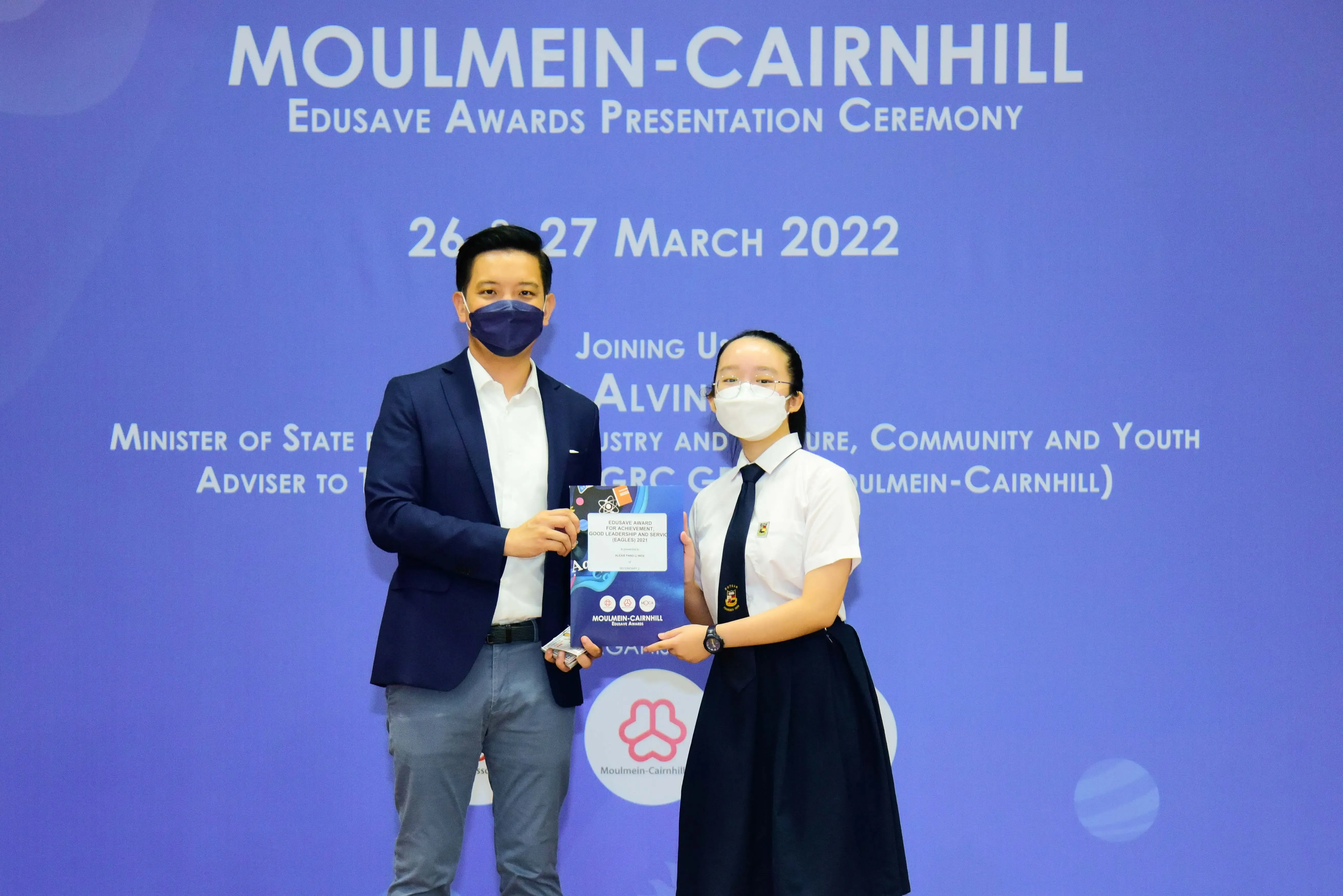 Two masked people hold an award at the Moulmein-Cairnhill Edusave Awards Presentation Ceremony, March 2022.