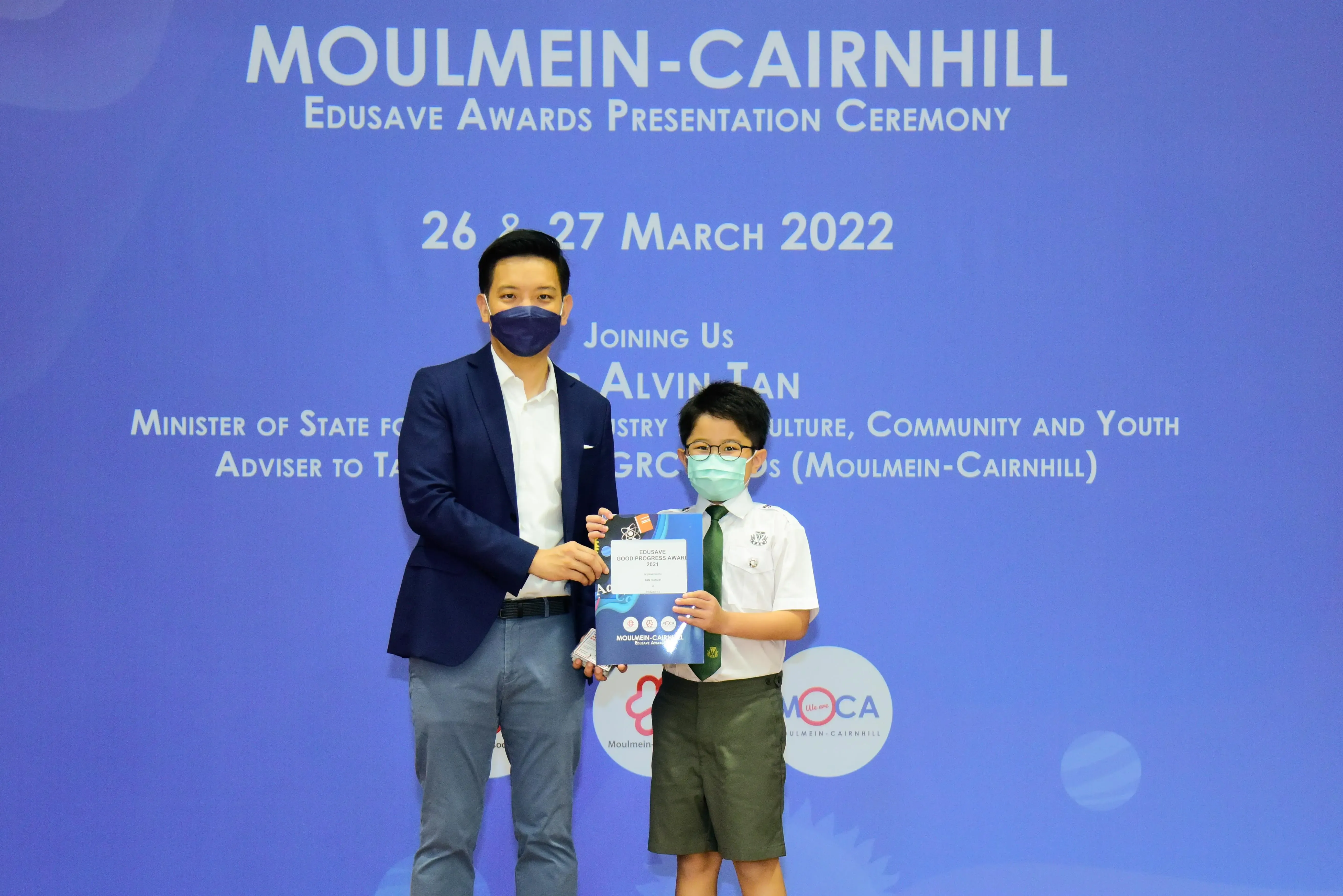 Man in suit and masked boy in school uniform hold an award certificate at Moulmein-Cairnhill Edusave ceremony, March 2022.