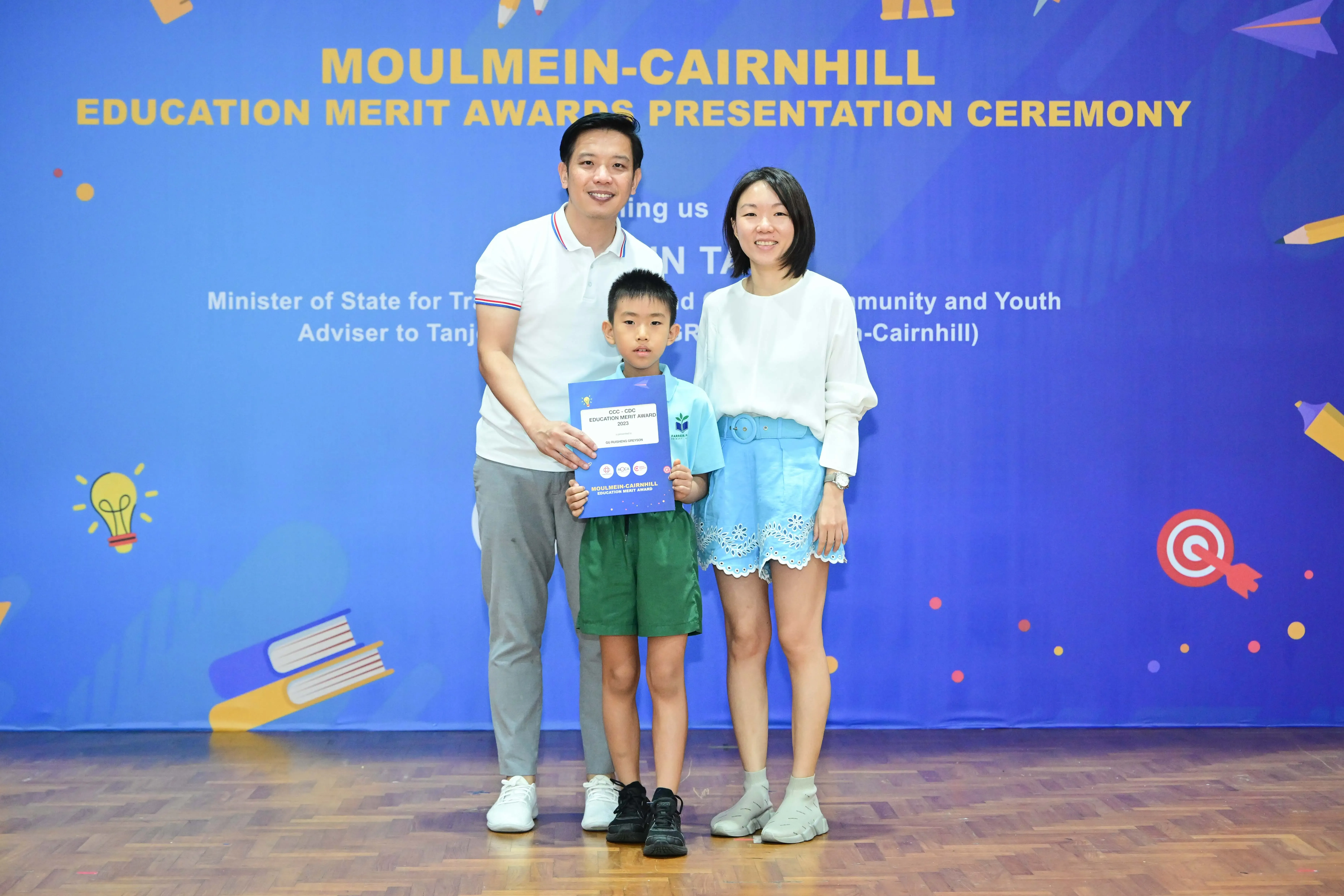 A boy holds an award certificate flanked by two adults at the Moulmein-Cairnhill Education Merit Awards ceremony.