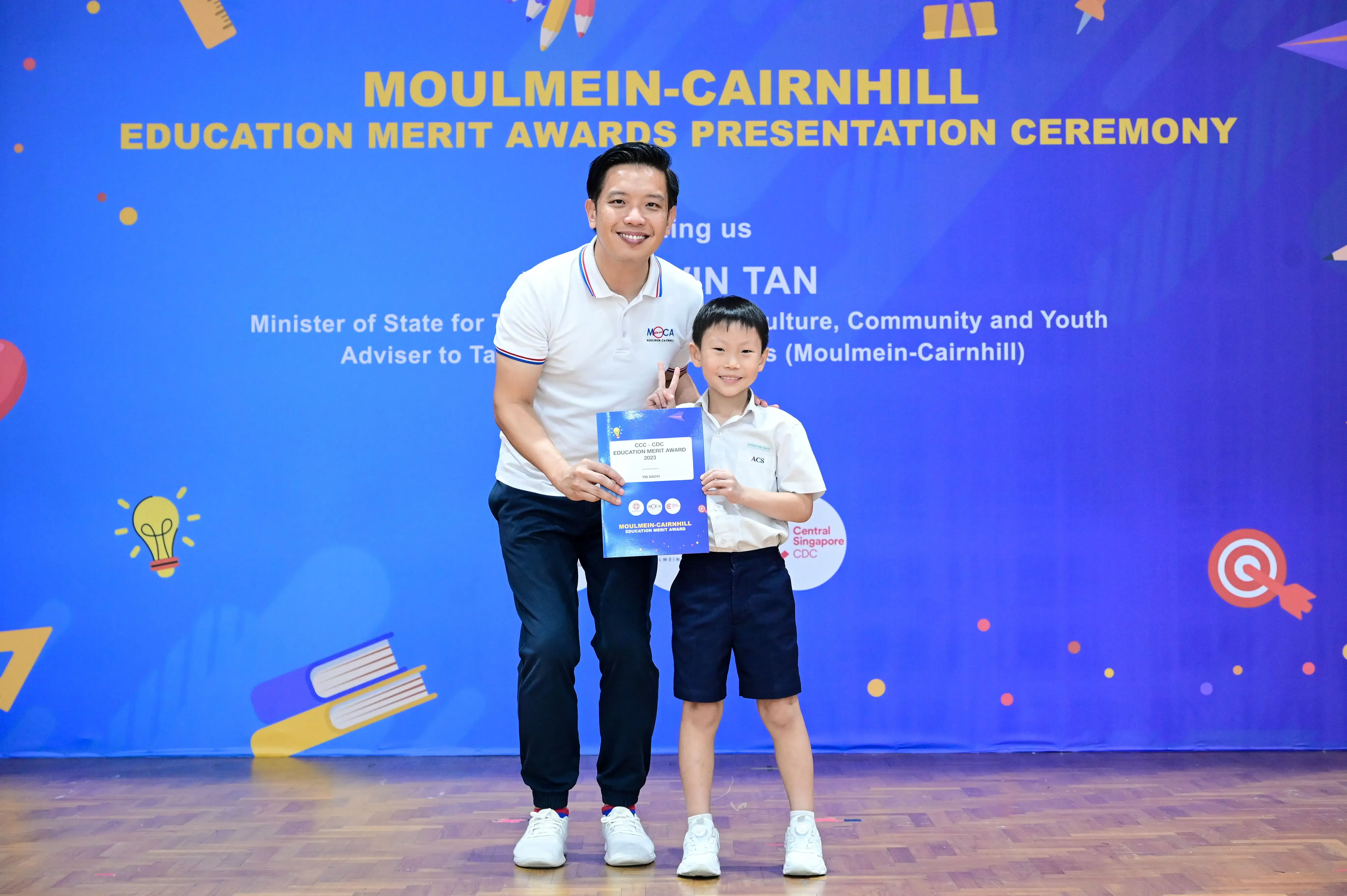 A man and a young boy in school uniform hold an award certificate at the Moulmein-Cairnhill Education Merit Awards ceremony.