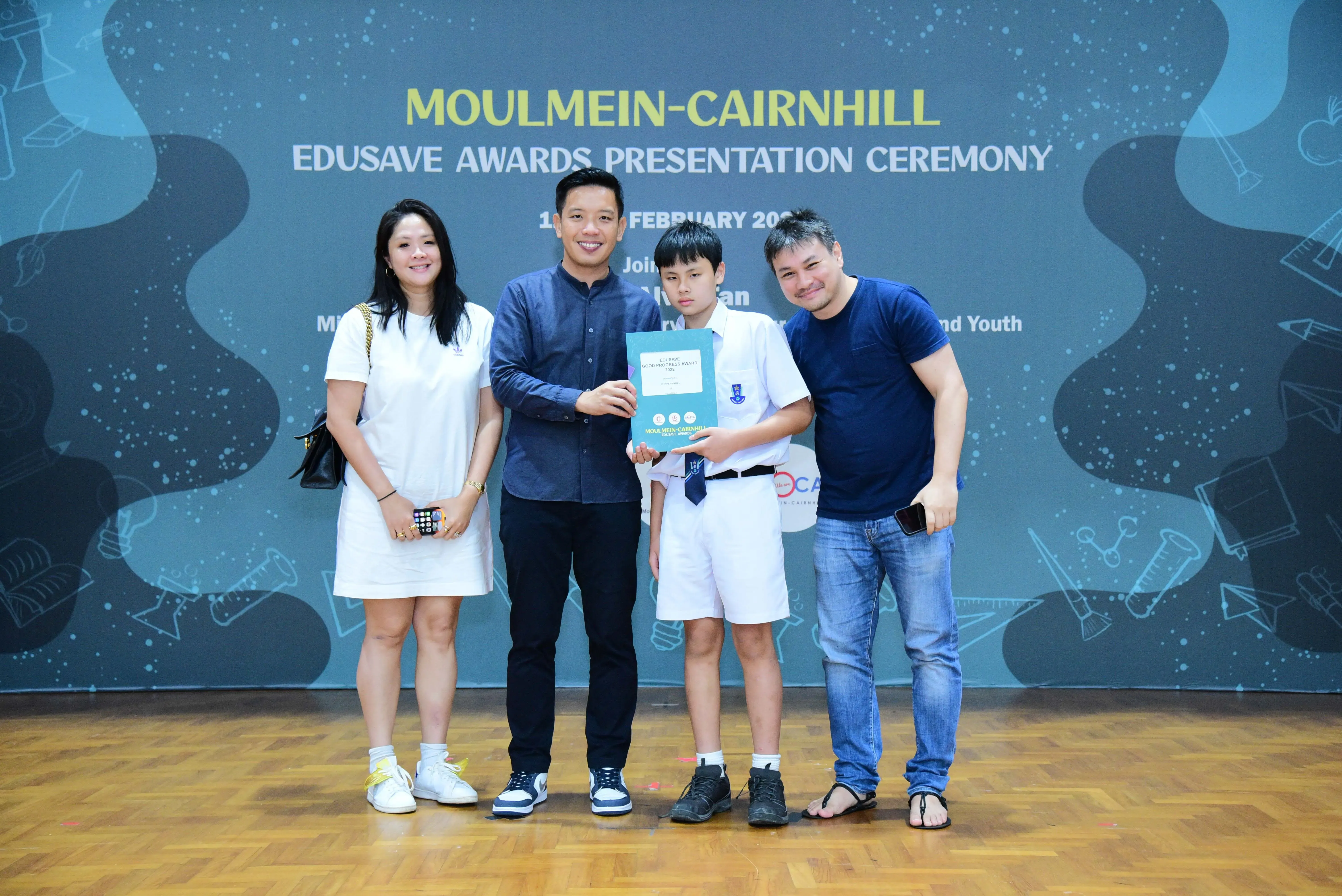 Four people pose at the Moulmein-Cairnhill Edusave Awards Presentation Ceremony, with a boy holding a certificate.