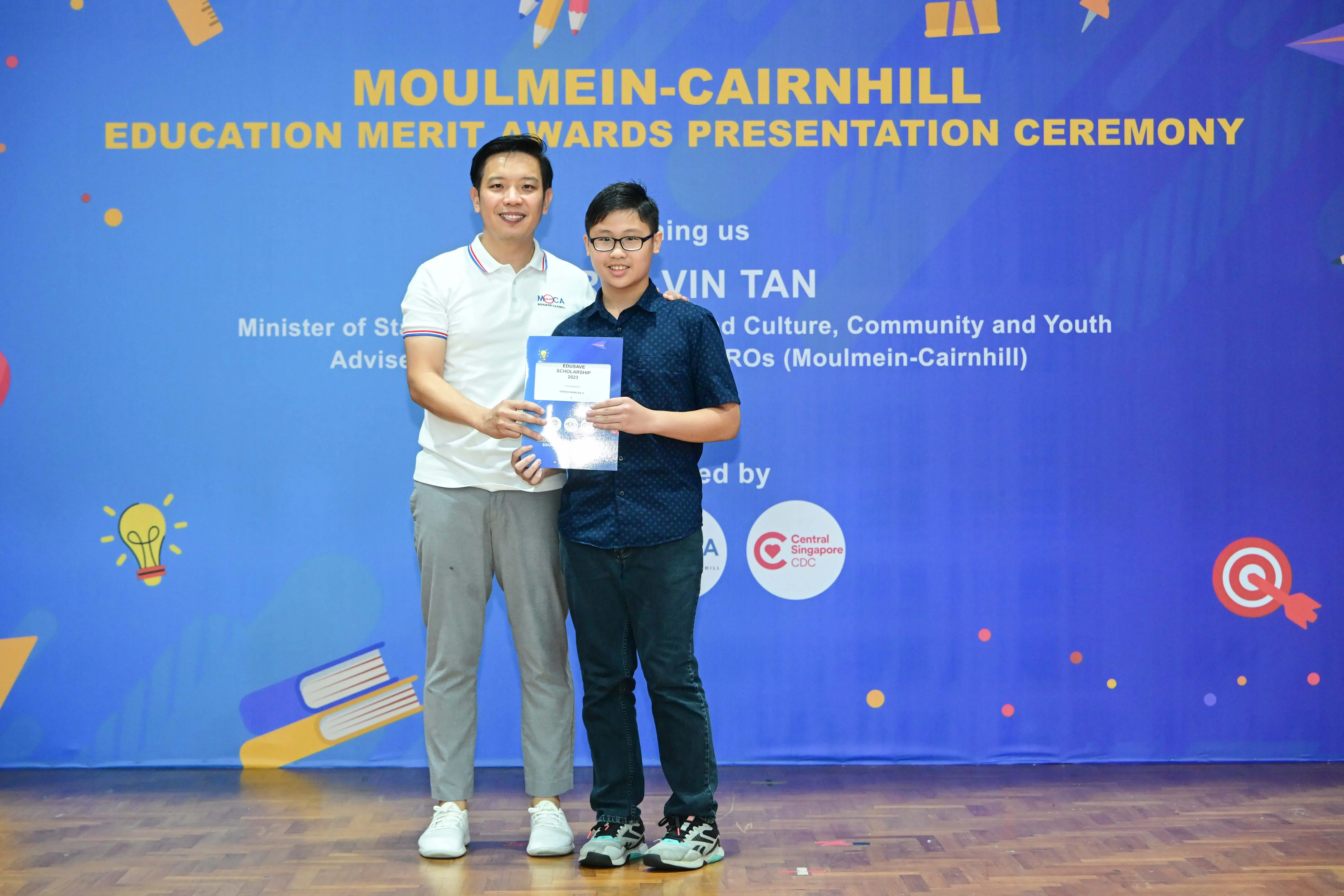 A man and a young boy hold a certificate together at the Moulmein-Cairnhill Education Merit Awards Presentation Ceremony.