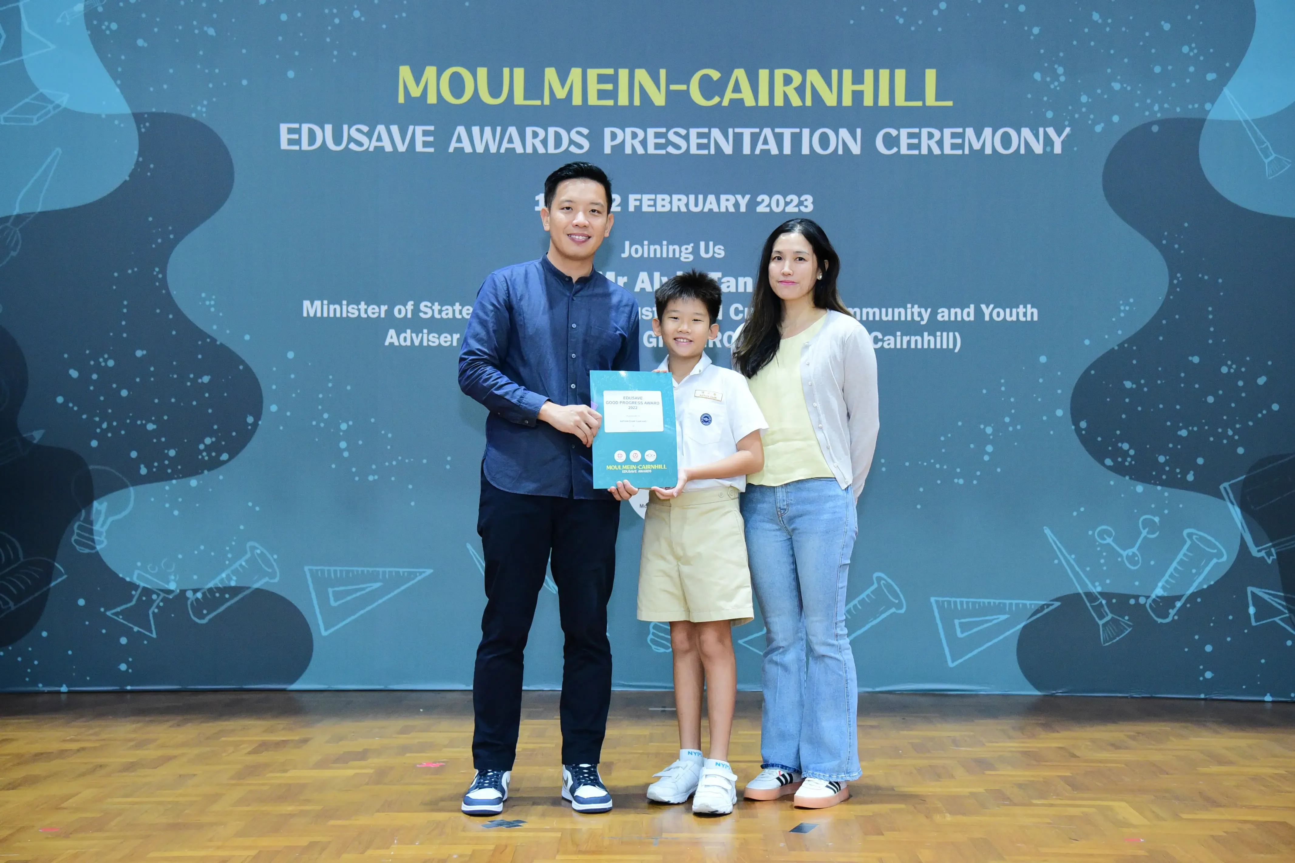A boy in school uniform holds a certificate with two adults at the Moulmein-Cairnhill Edusave Awards Ceremony 2023.