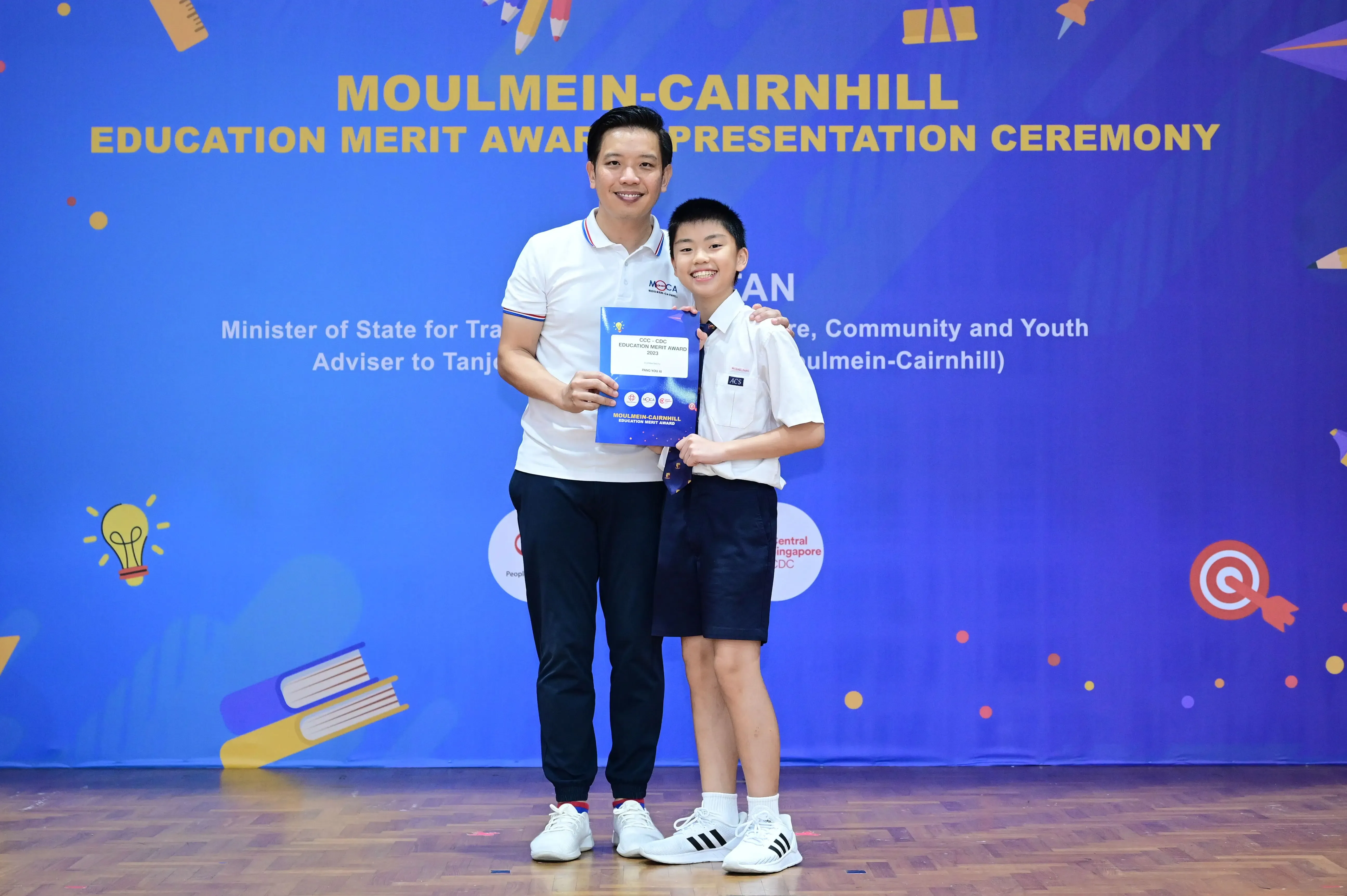 A man and a young boy pose holding an award booklet at the Moulmein-Cairnhill Education Merit Award Ceremony.