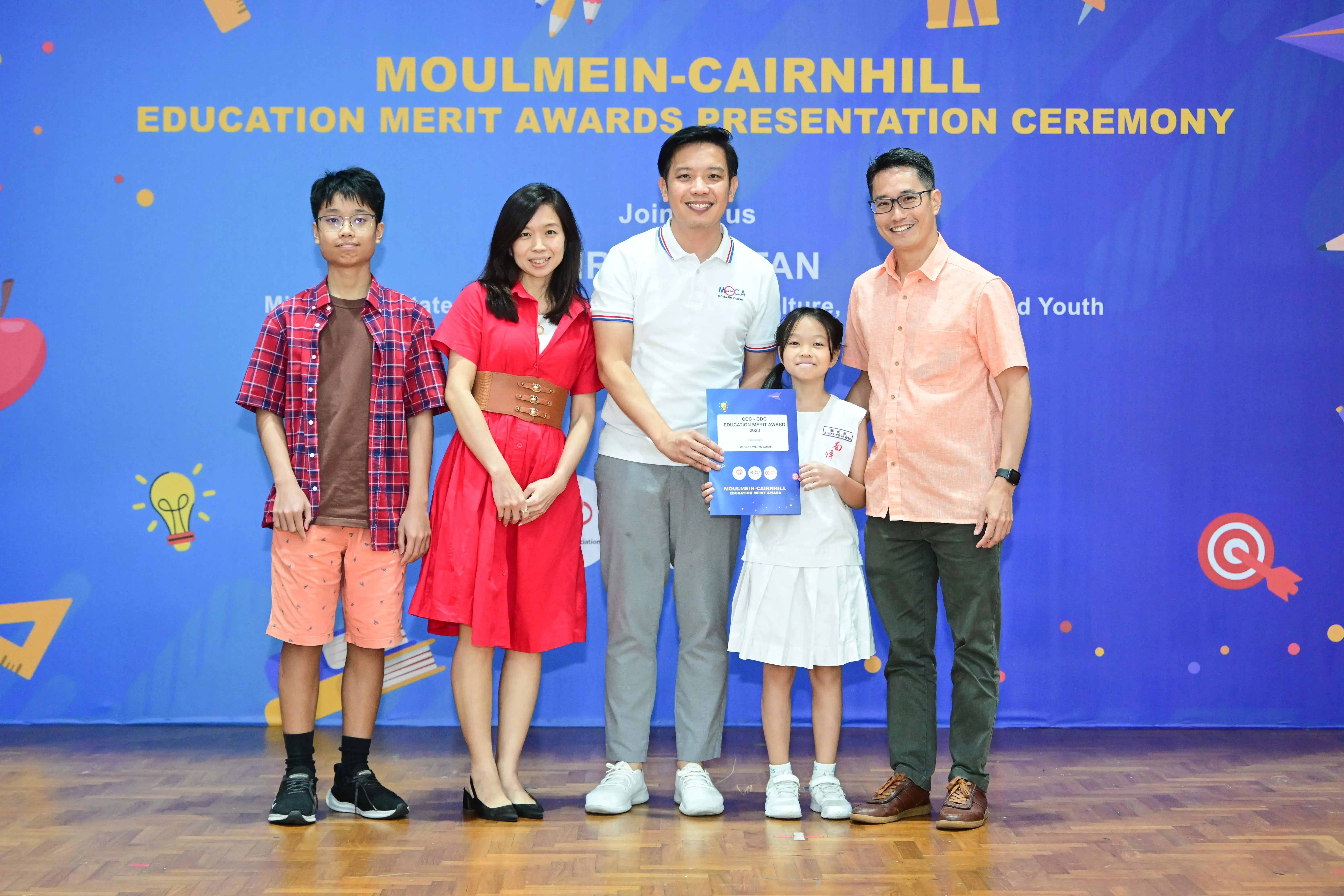 Five people pose at the Moulmein-Cairnhill Education Merit Awards Presentation Ceremony, with a girl holding a certificate.