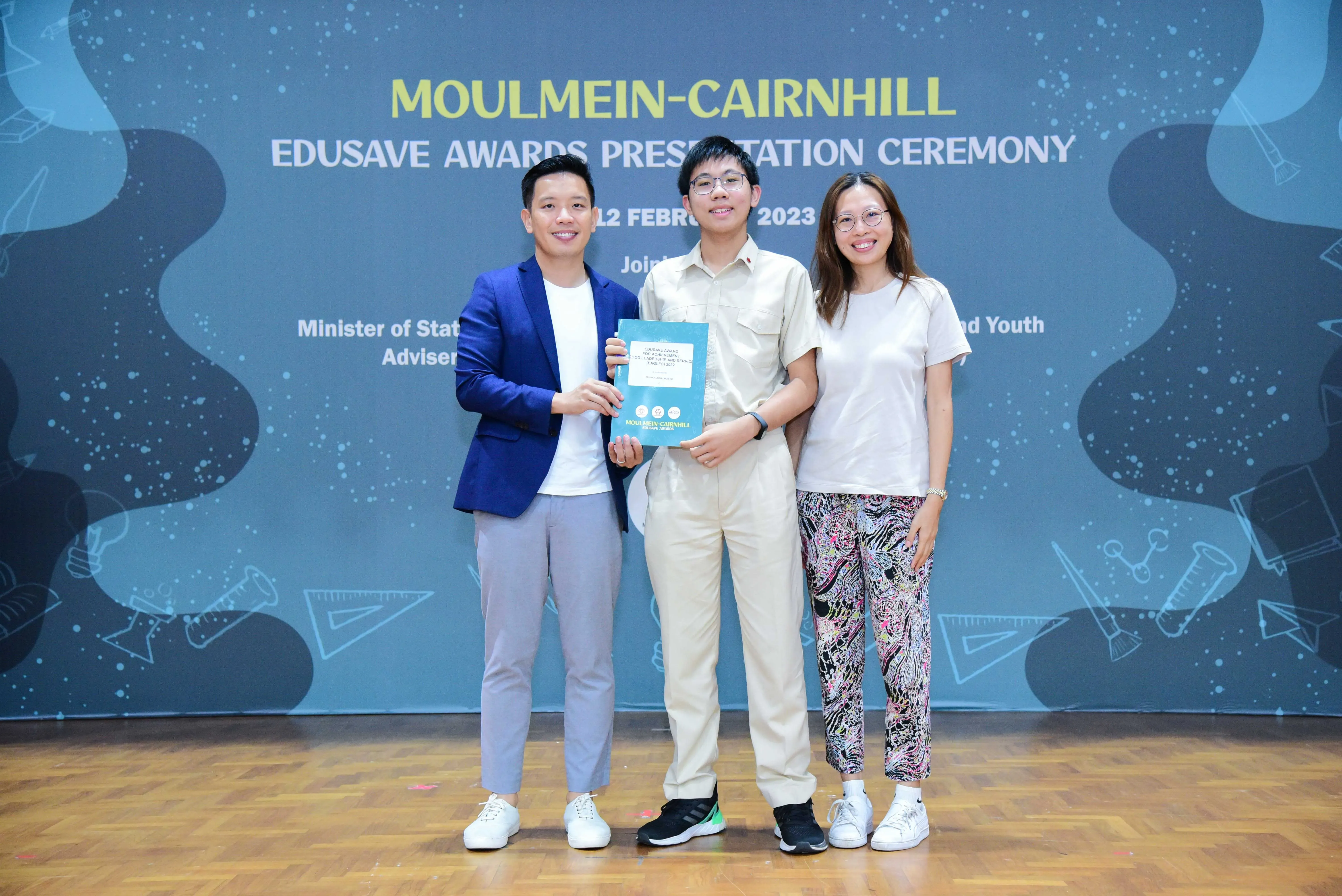 Three people smile on stage at the Moulmein-Cairnhill Edusave Awards Presentation Ceremony 2023, center person holds a certificate.