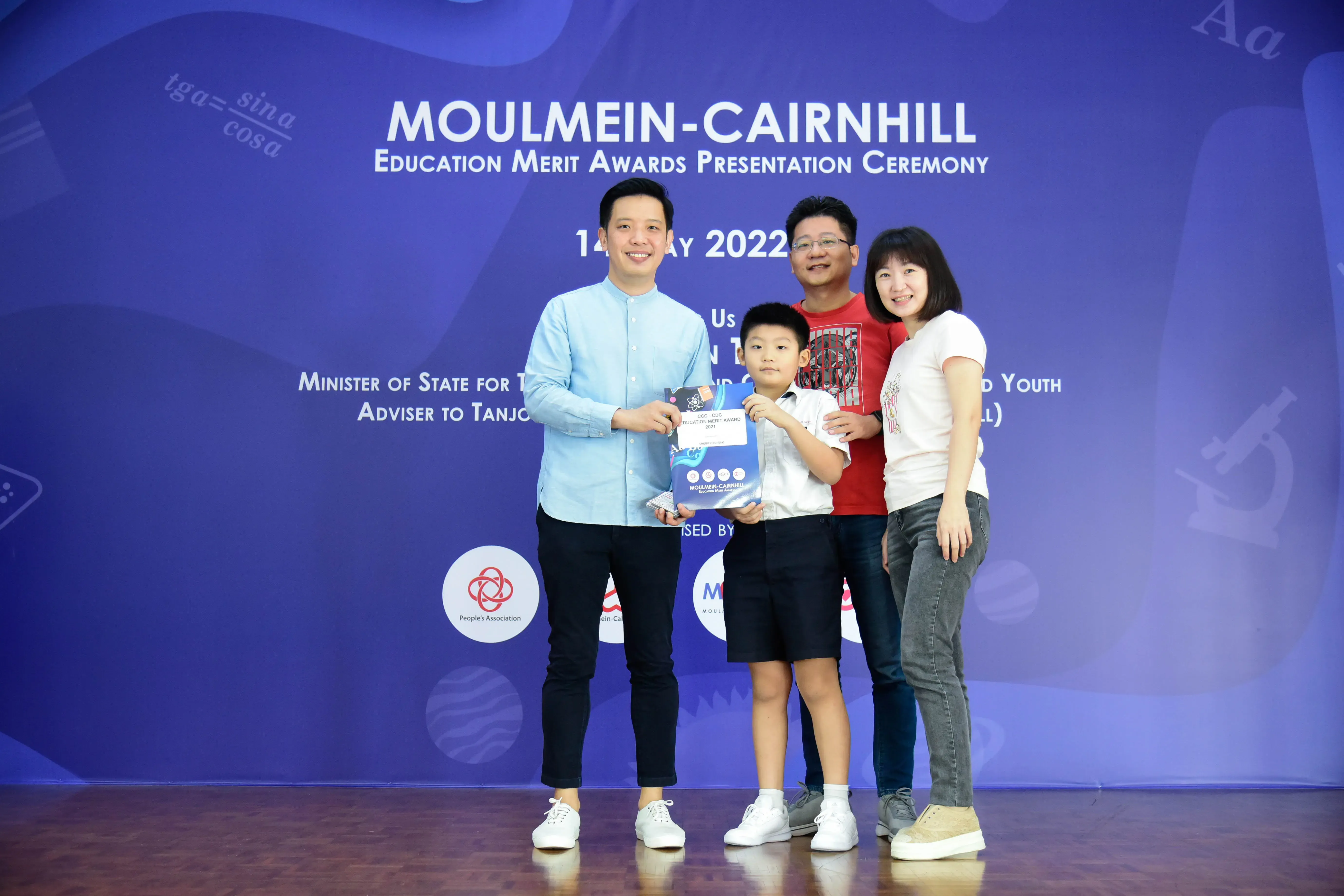 Four people pose at the Moulmein-Cairnhill Education Merit Awards Ceremony, with a boy holding an award certificate.