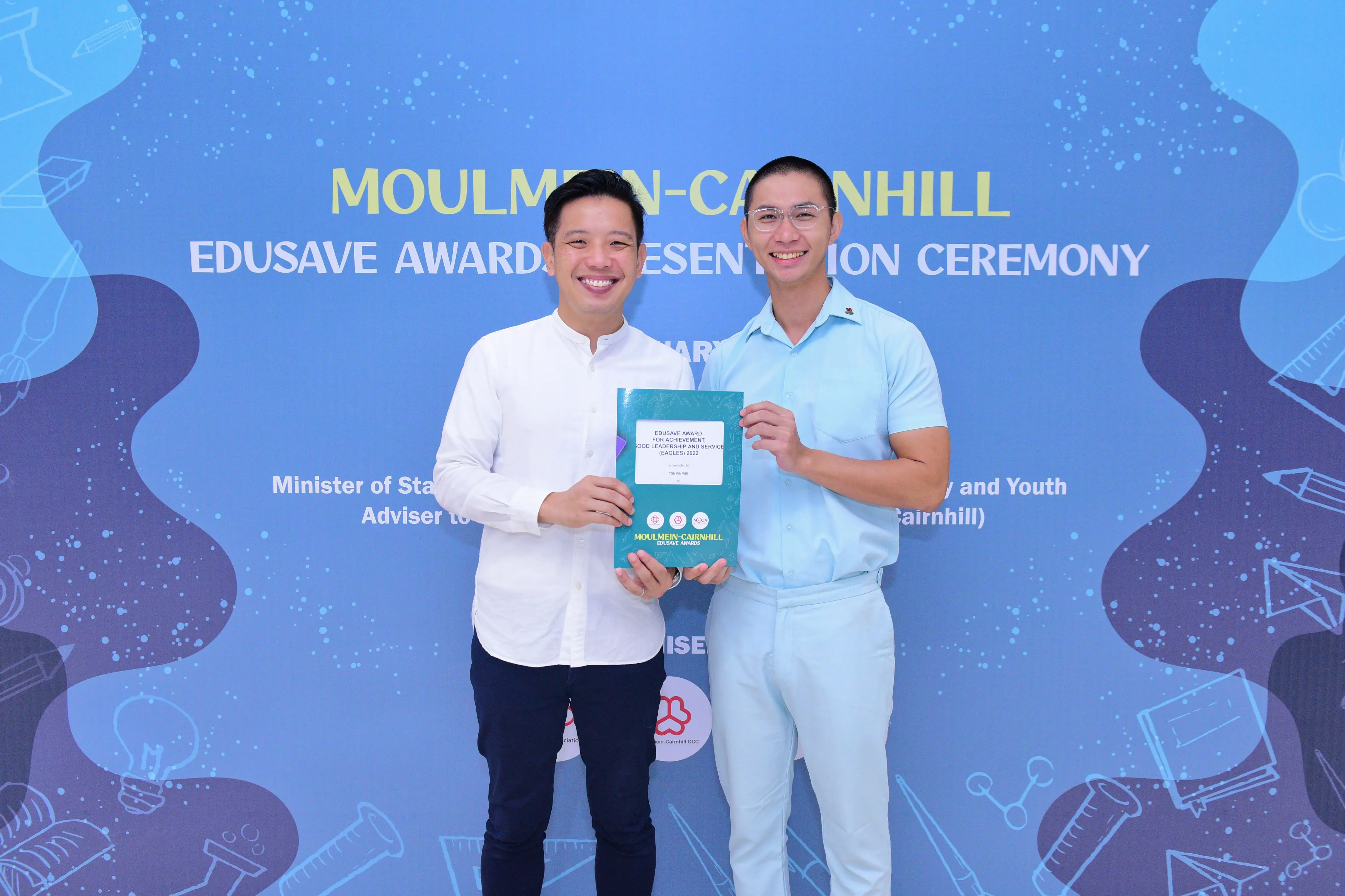 Two smiling men hold an Edusave award booklet at the Moulmein-Cairnhill Edusave Awards Presentation Ceremony.