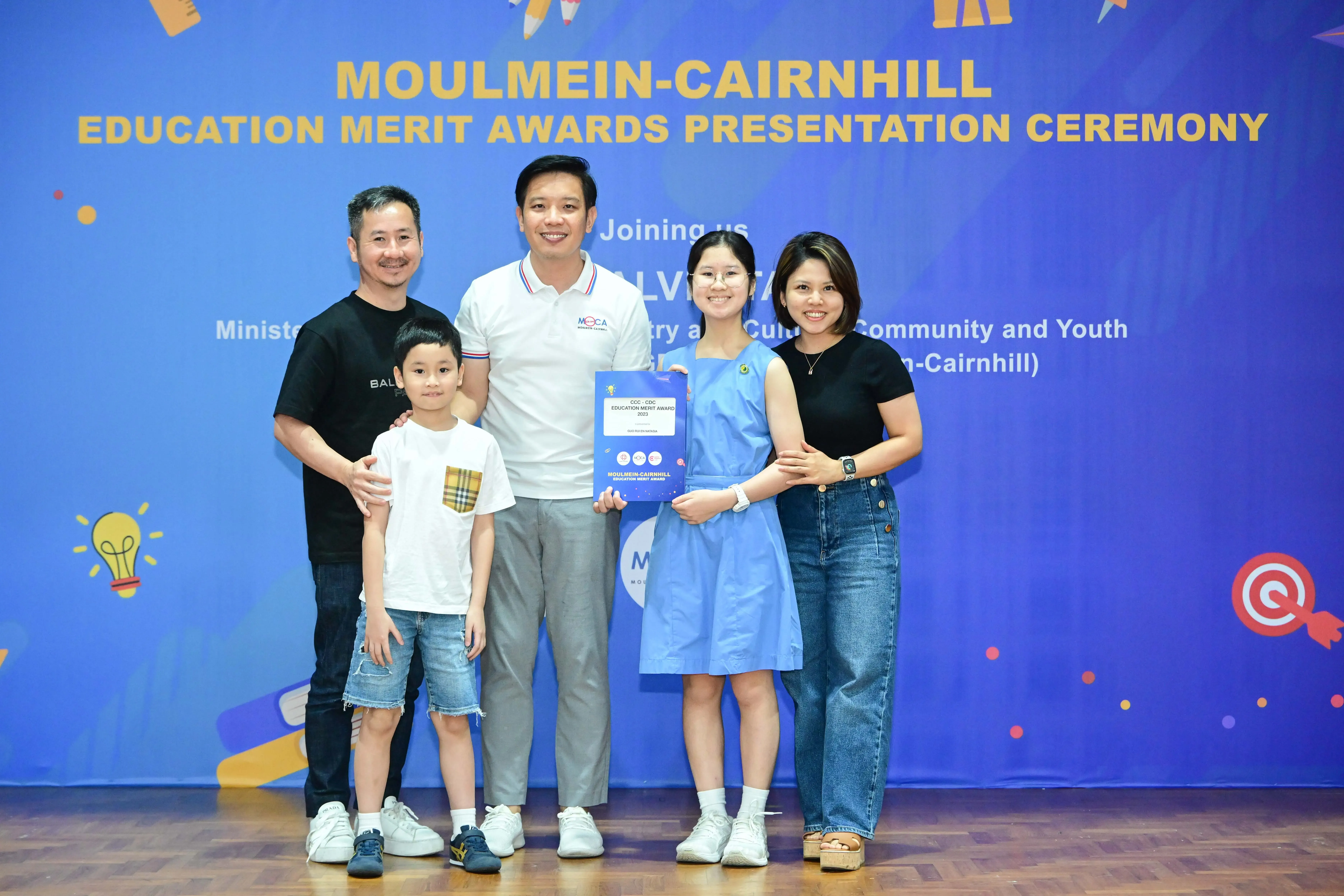 Four adults and a child pose at the Moulmein-Cairnhill Education Merit Awards Presentation Ceremony, with a girl holding an award.