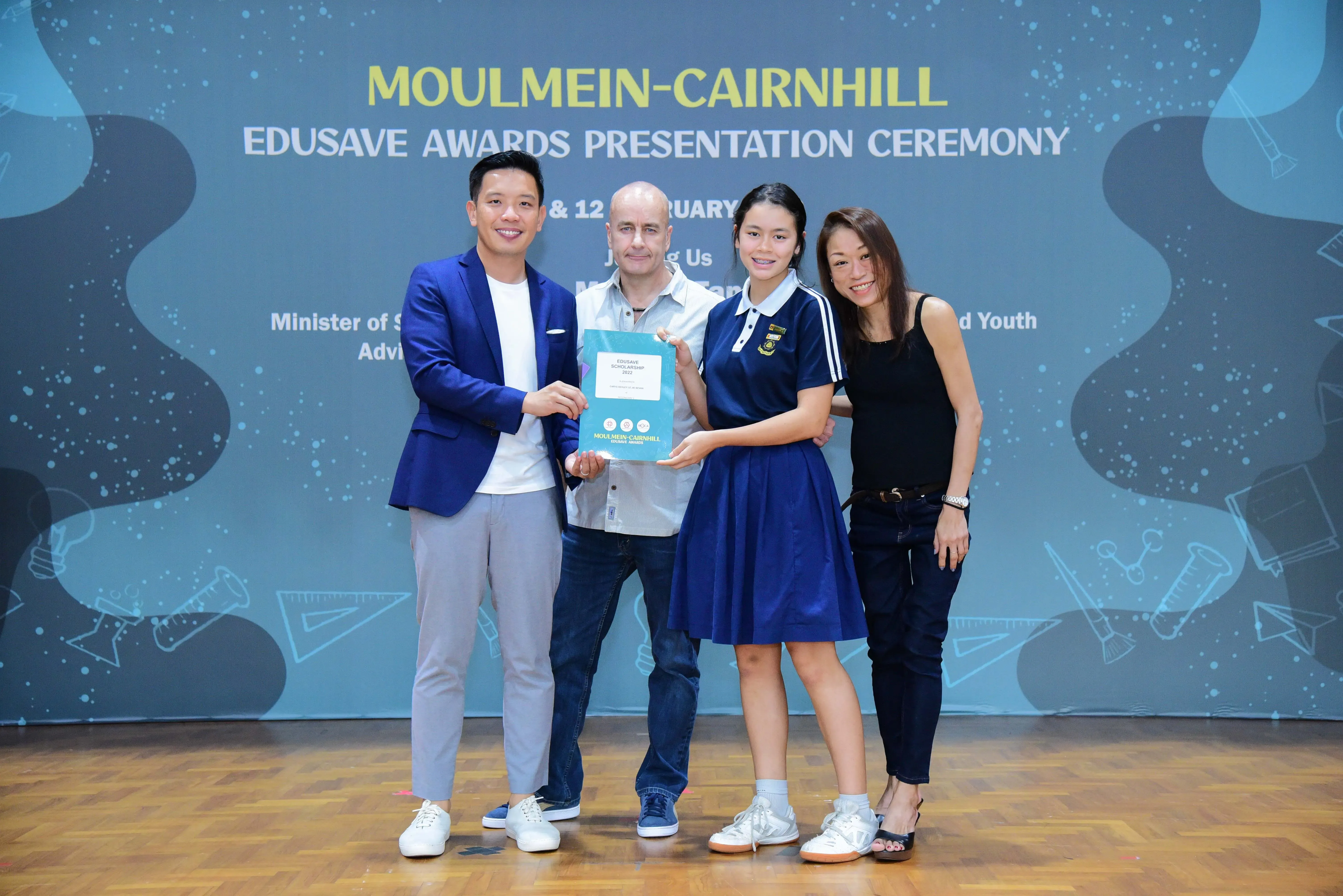Four people pose holding a certificate at the Moulmein-Cairnhill Edusave Awards Presentation Ceremony backdrop.