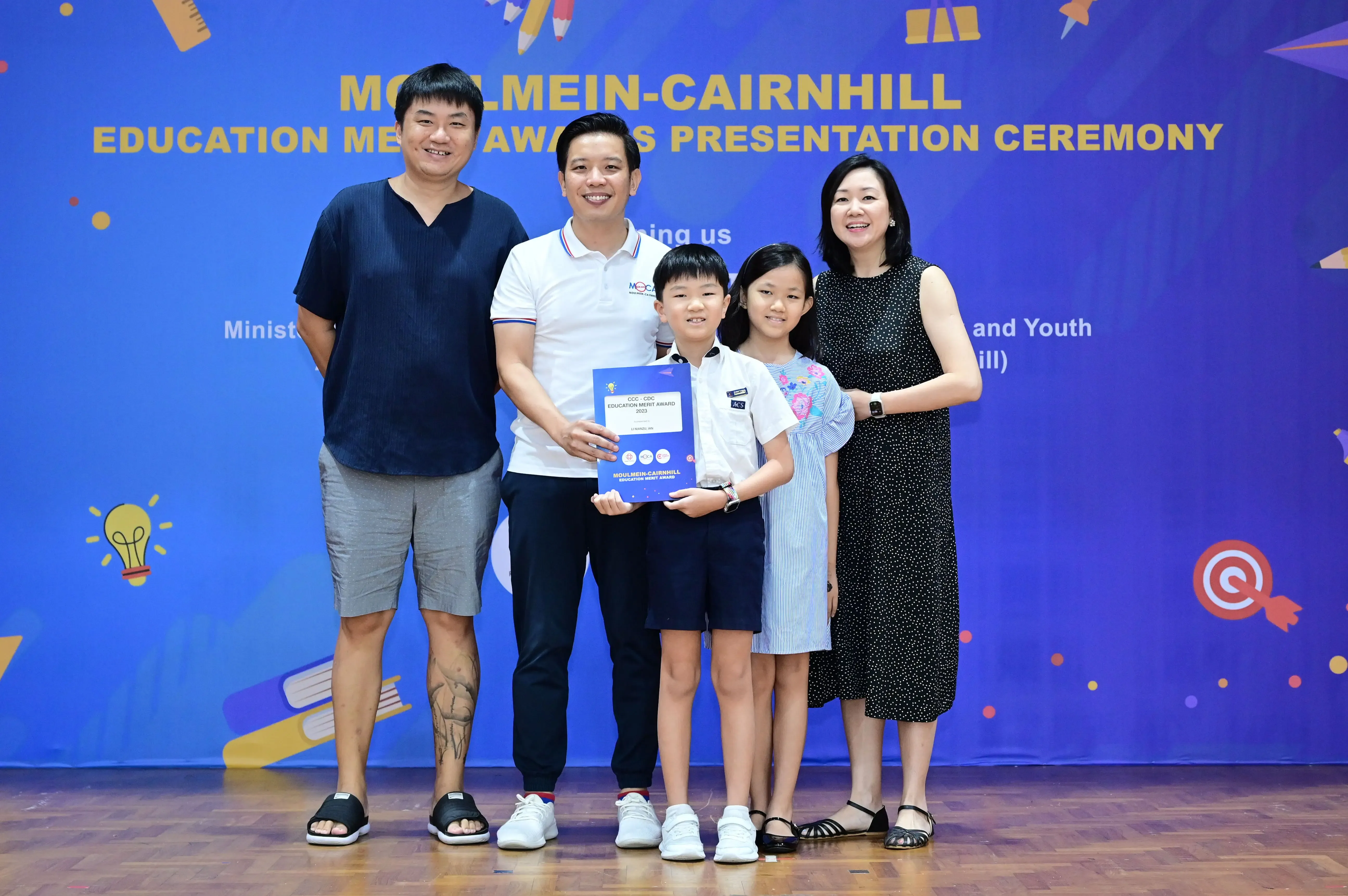 Five people pose before a Moulmein-Cairnhill Education Merit Awards banner, two children holding a blue award certificate.
