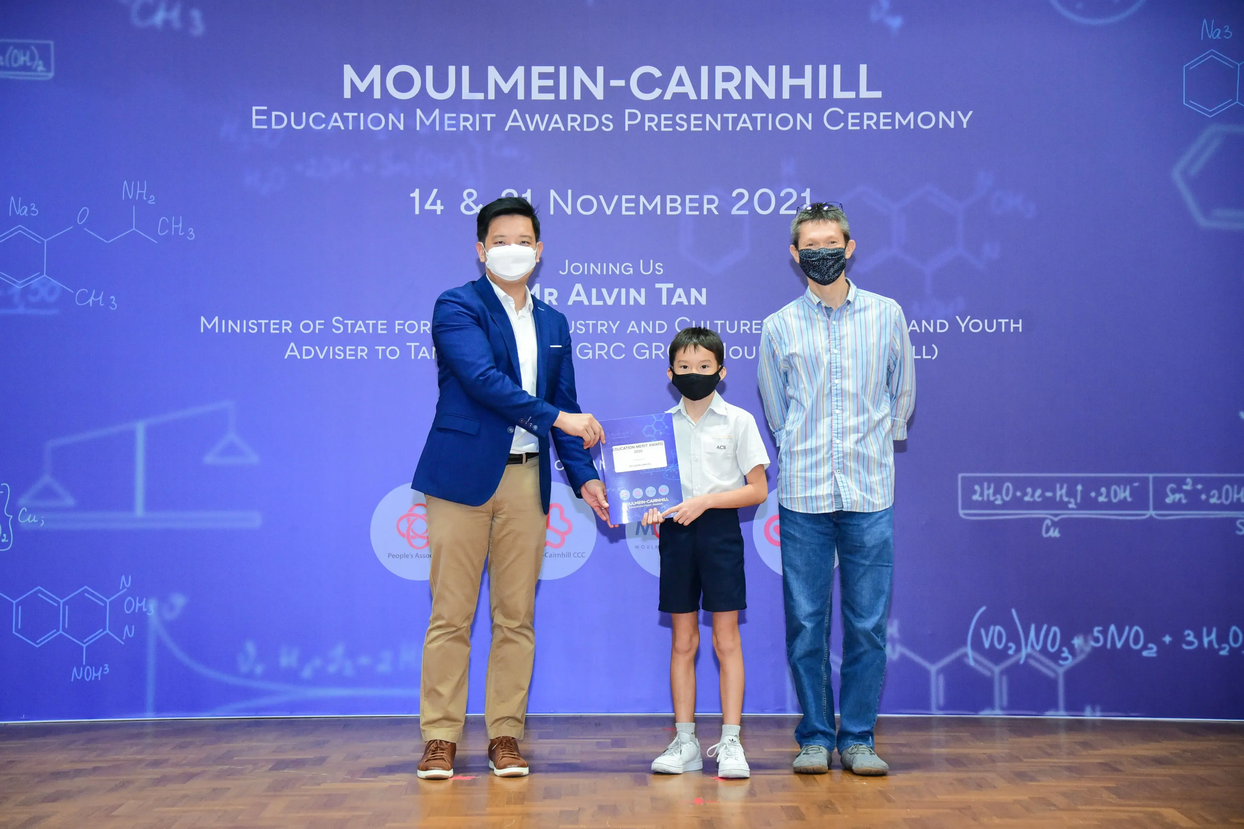 Two masked adults and a masked child hold an award at the Moulmein-Cairnhill Education Merit Awards Ceremony 2021.