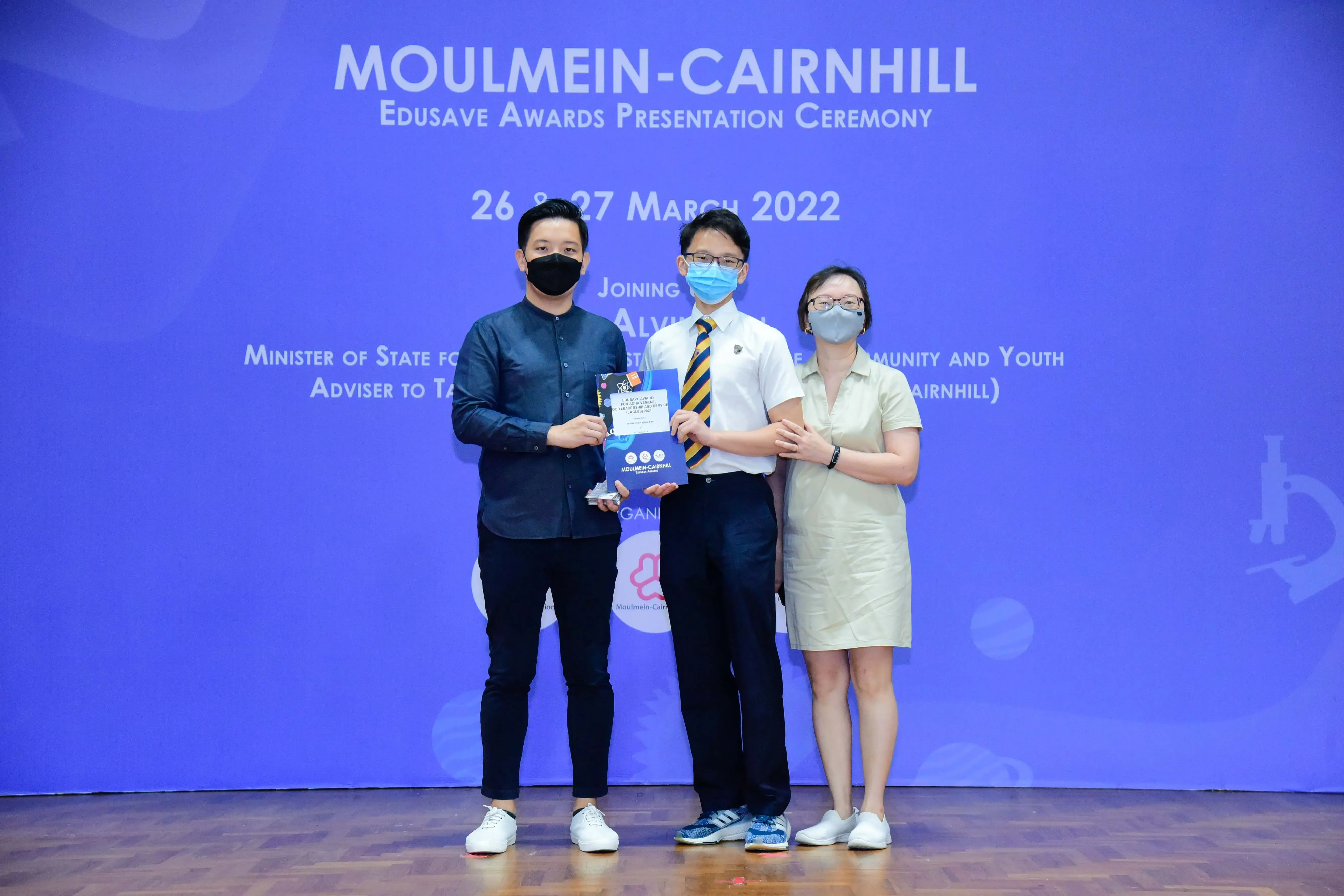Three masked people stand on a stage holding an award at the Moulmein-Cairnhill Edusave Awards Ceremony, March 2022.