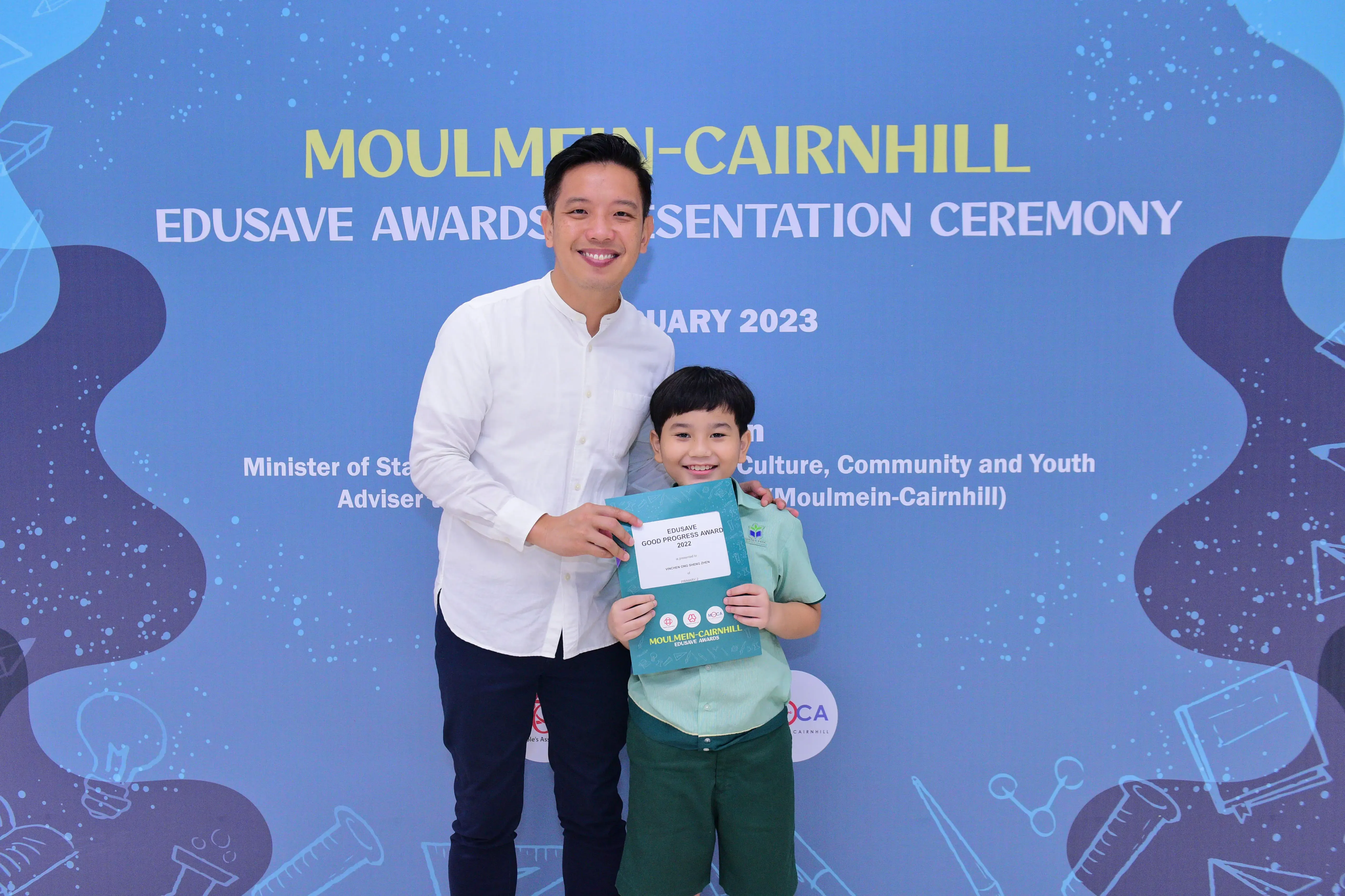A man and a young boy smile holding an Edusave award at the Moulmein-Cairnhill Awards Presentation Ceremony 2023.