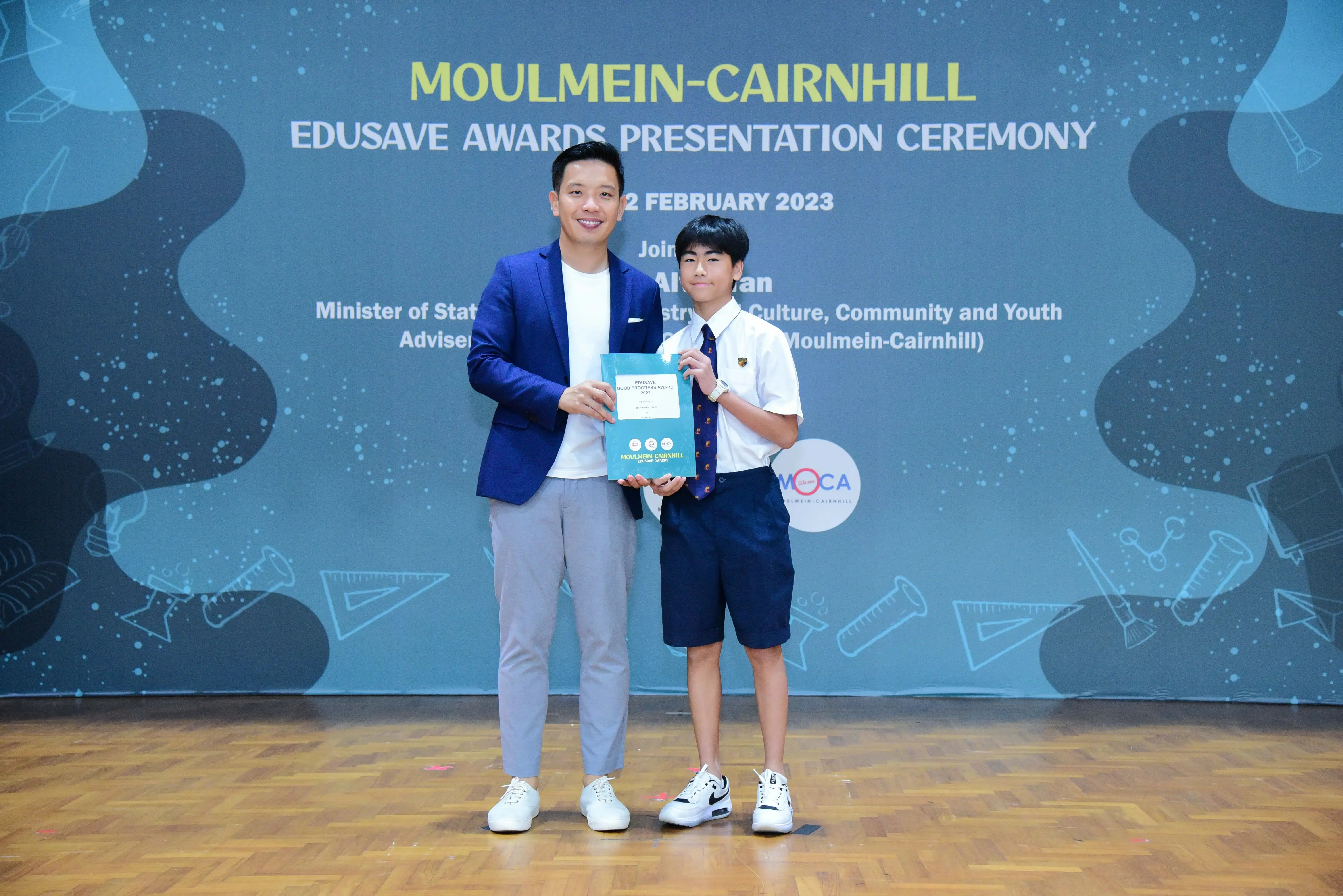 An adult in a blue blazer and a uniformed student hold an award at the Moulmein-Cairnhill Edusave Awards Ceremony 2023.