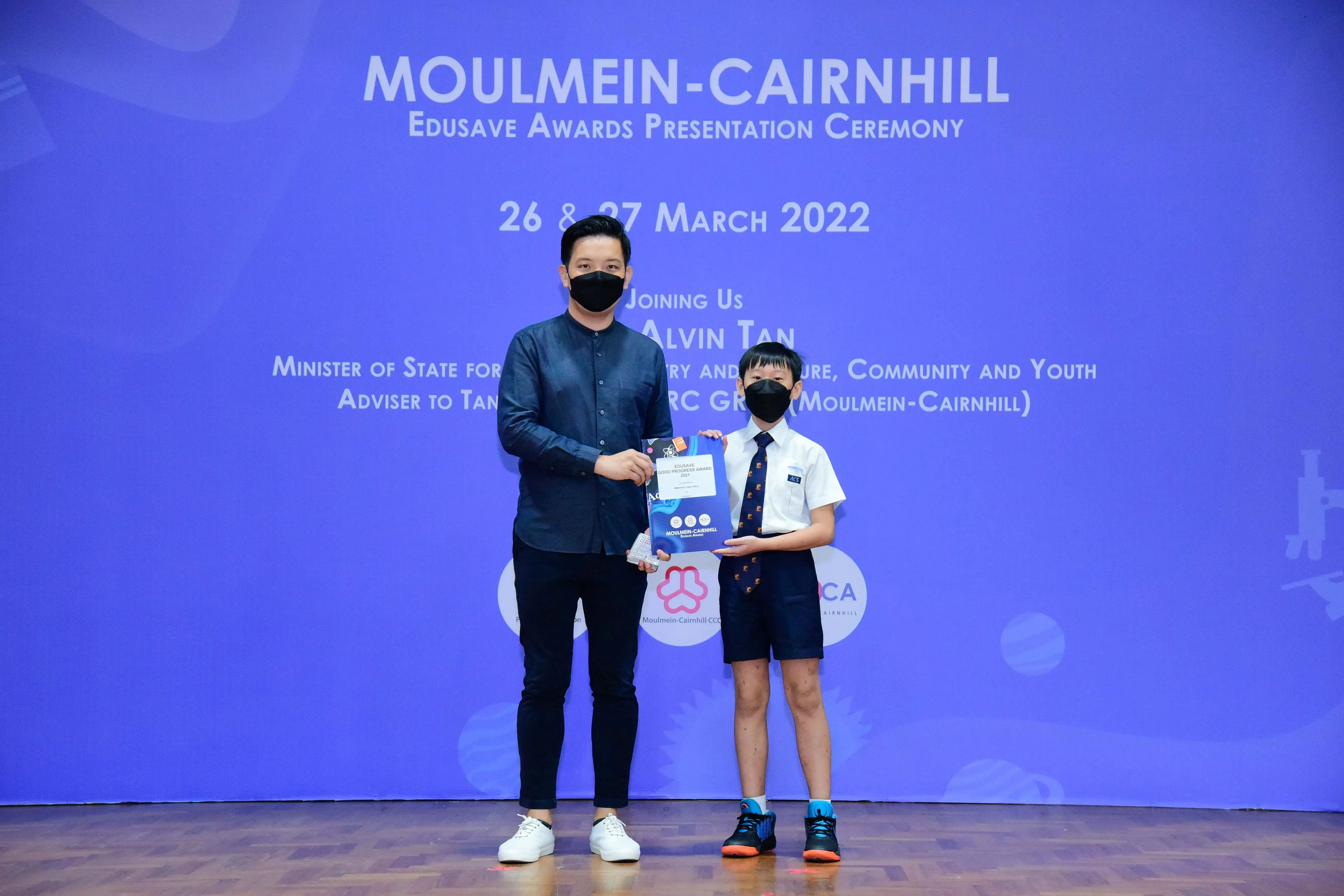 An adult and a young boy in masks hold an award certificate at the Moulmein-Cairnhill Edusave Awards Ceremony 2022.