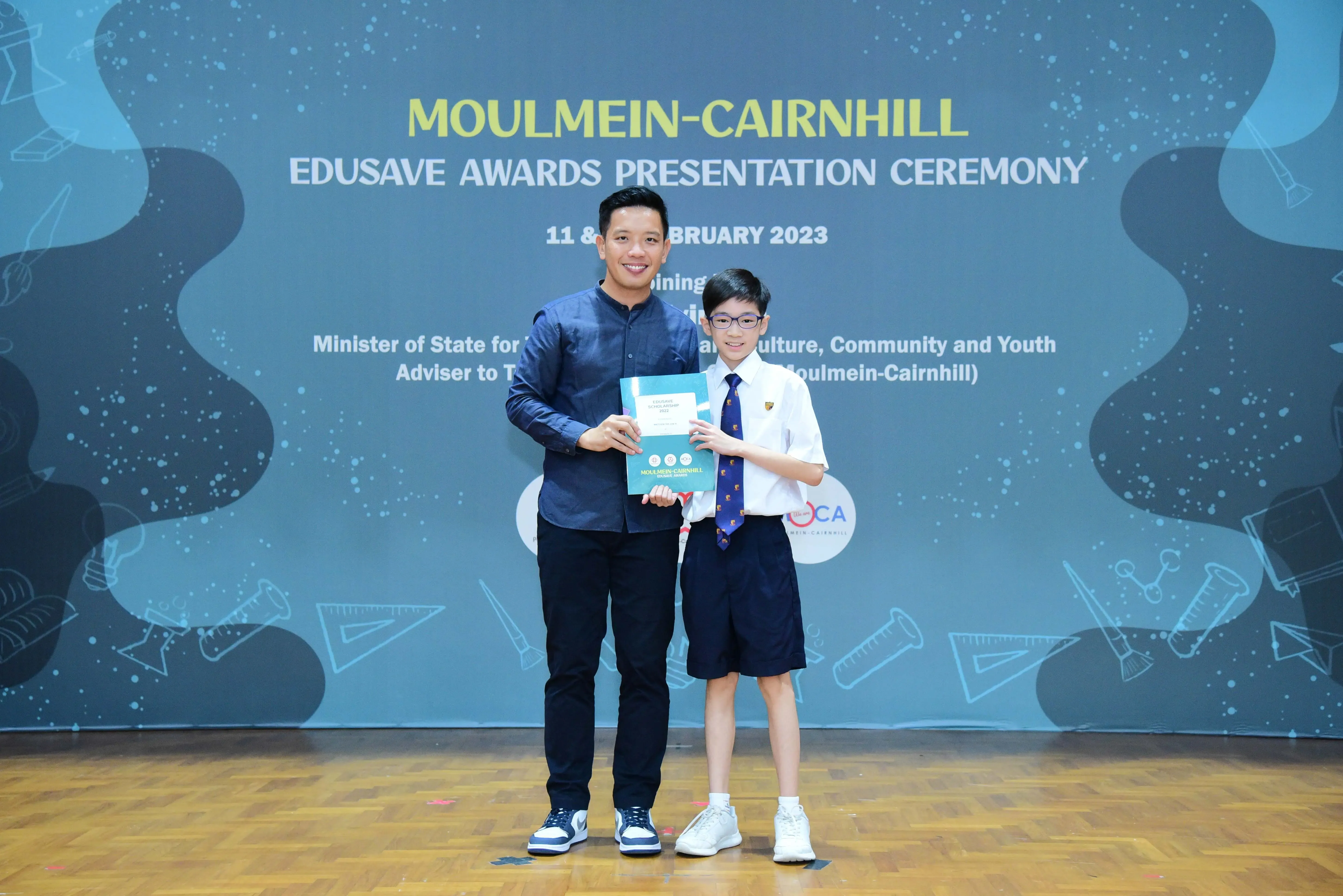 An adult and a young student in uniform hold a certificate on stage at the Moulmein-Cairnhill Edusave Awards Ceremony 2023.