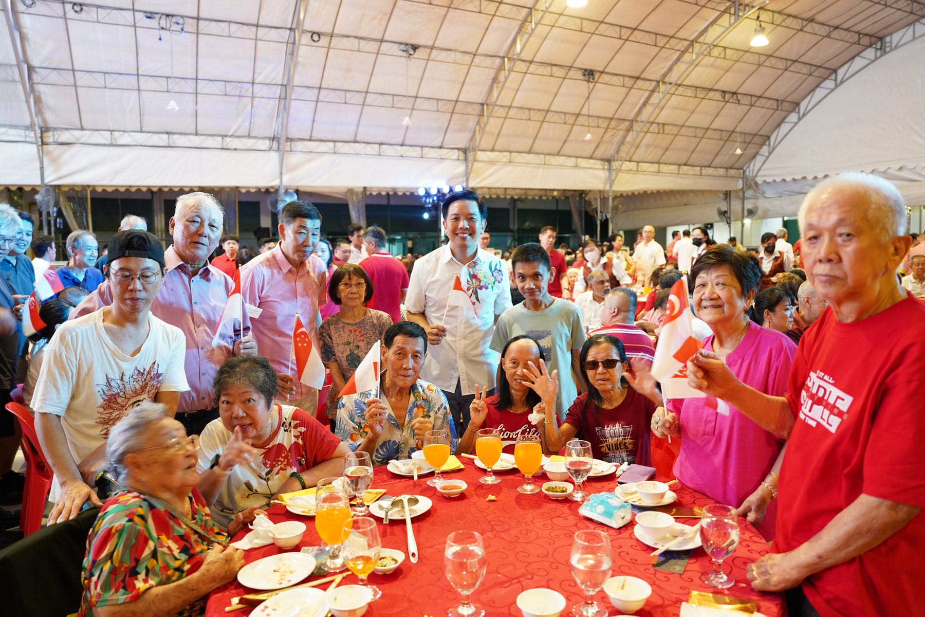 A large group of people in red and white clothes gather at a banquet table under a tent, waving small flags.