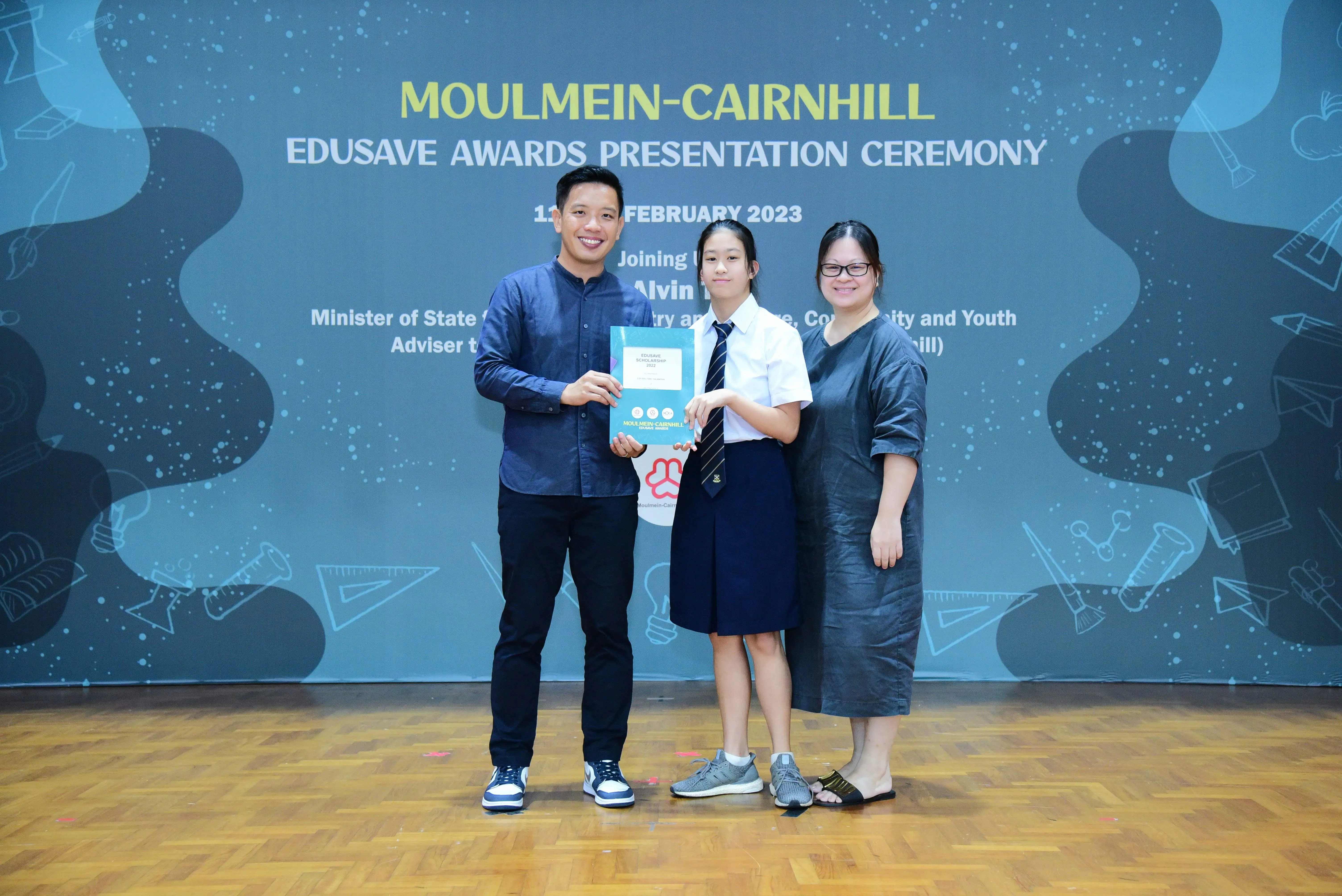 Three people pose together holding an award booklet at the Moulmein-Cairnhill Edusave Awards Presentation Ceremony 2023.