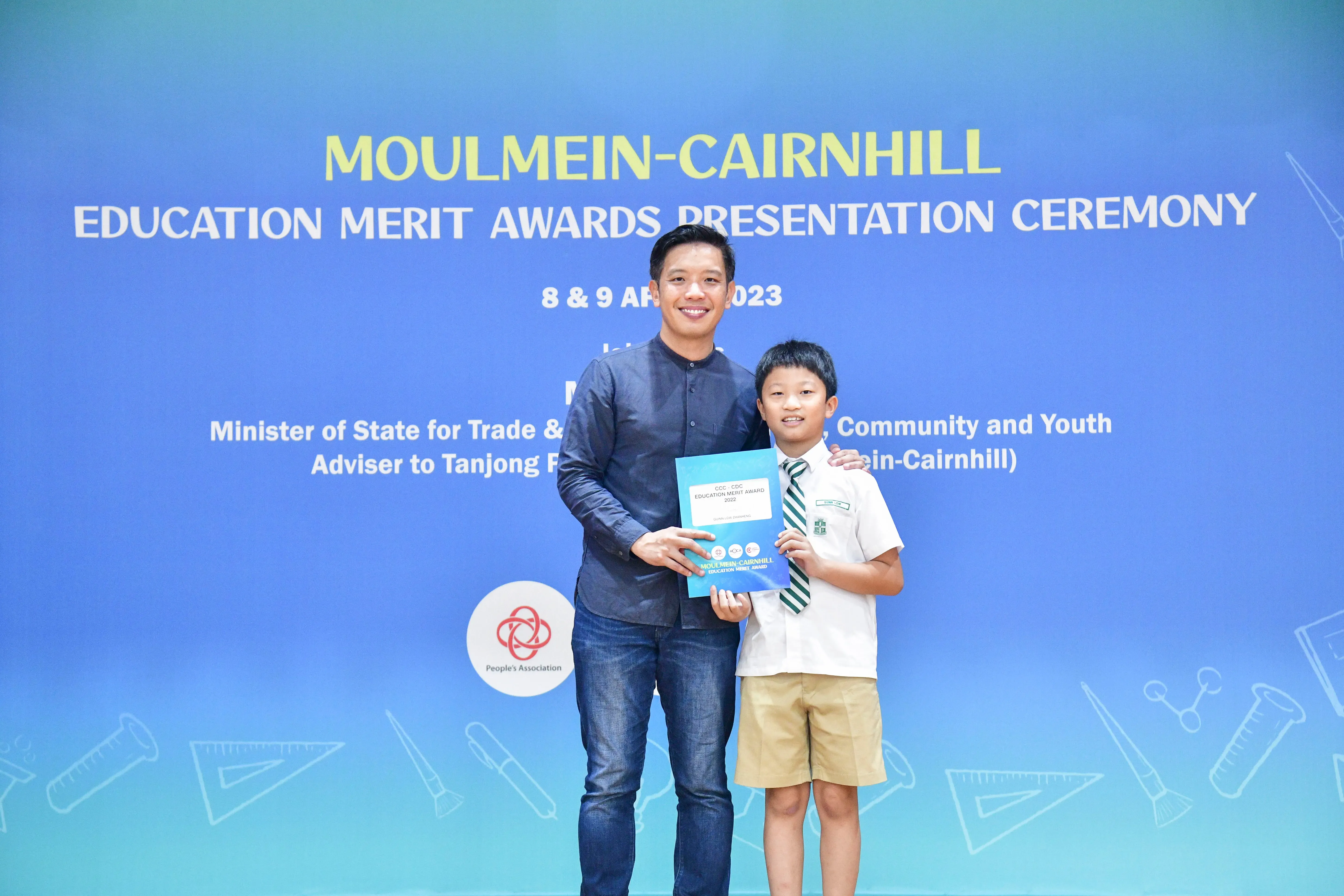 An adult and a young boy hold an award certificate in front of a Moulmein-Cairnhill Education Merit Awards banner.