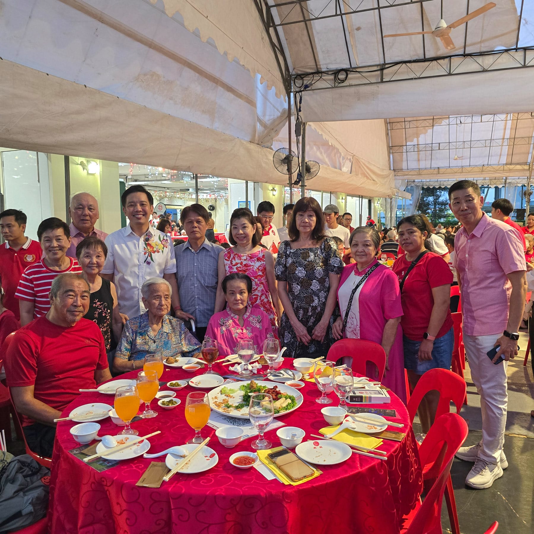 A group of people posing around a red banquet table with food and drinks at an outdoor celebration event.