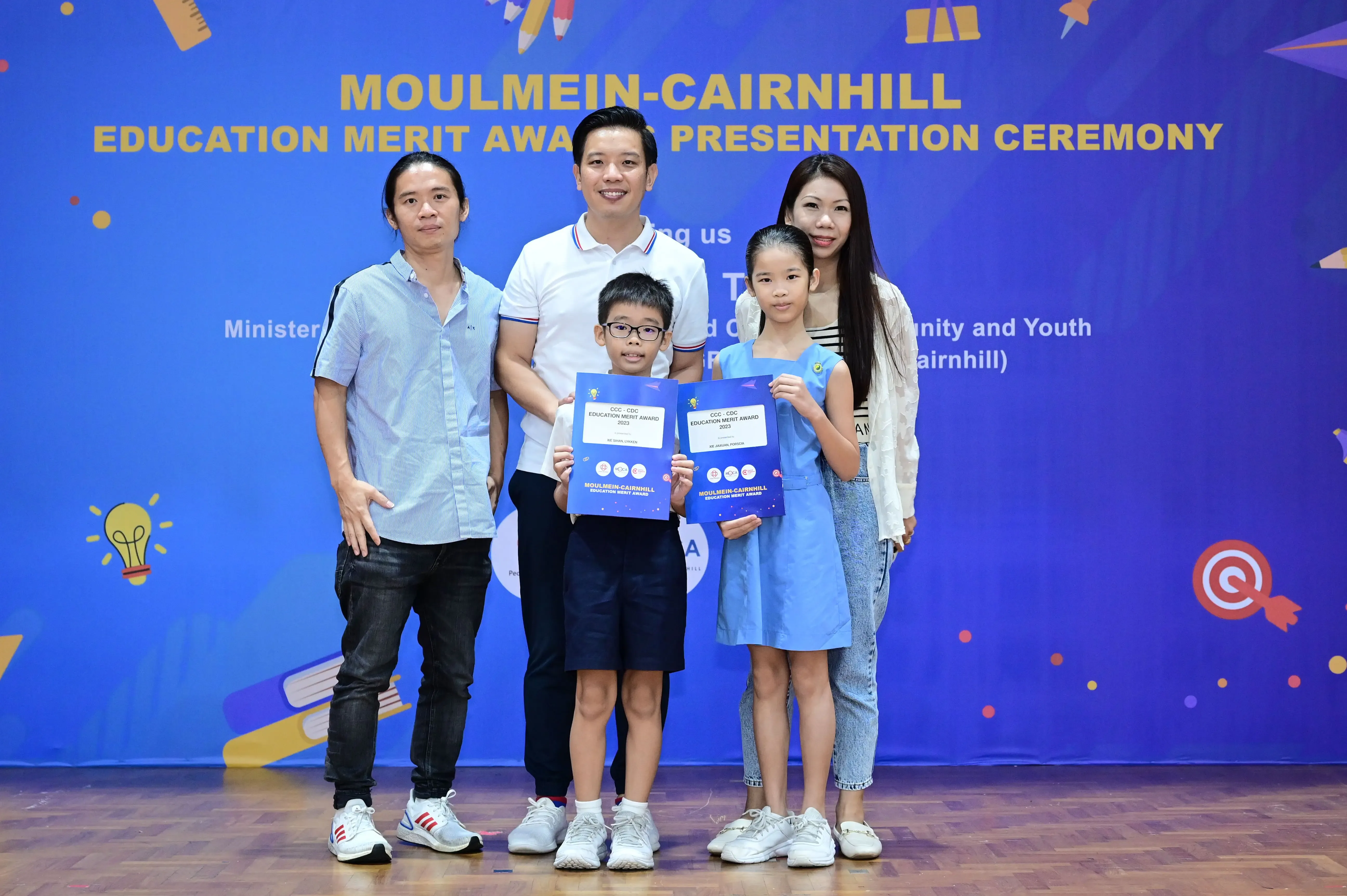 Two children holding merit award certificates pose with three adults at the Moulmein-Cairnhill Education Merit Awards ceremony.