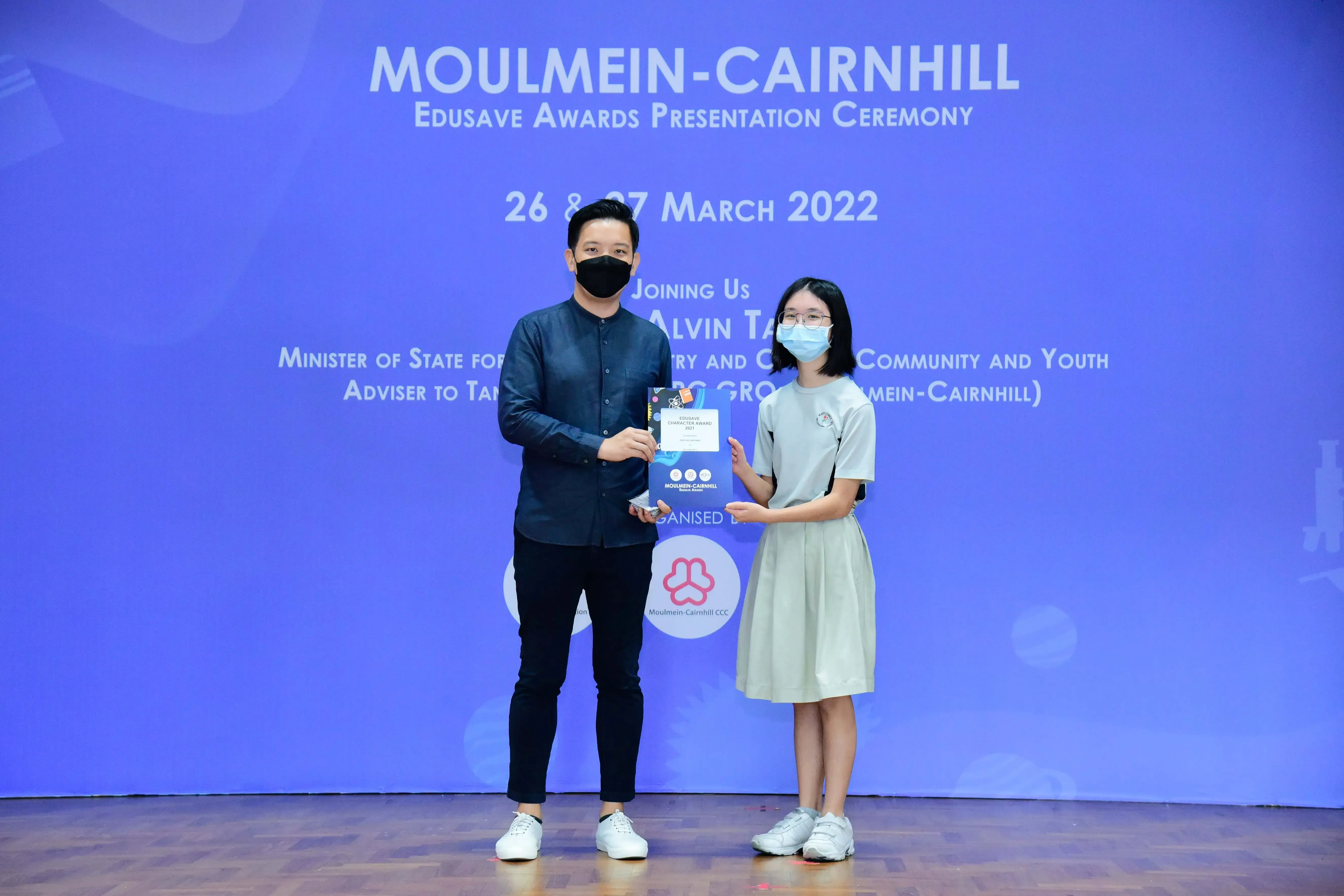Two masked people hold an award at the Moulmein-Cairnhill Edusave Awards Presentation Ceremony, March 2022.