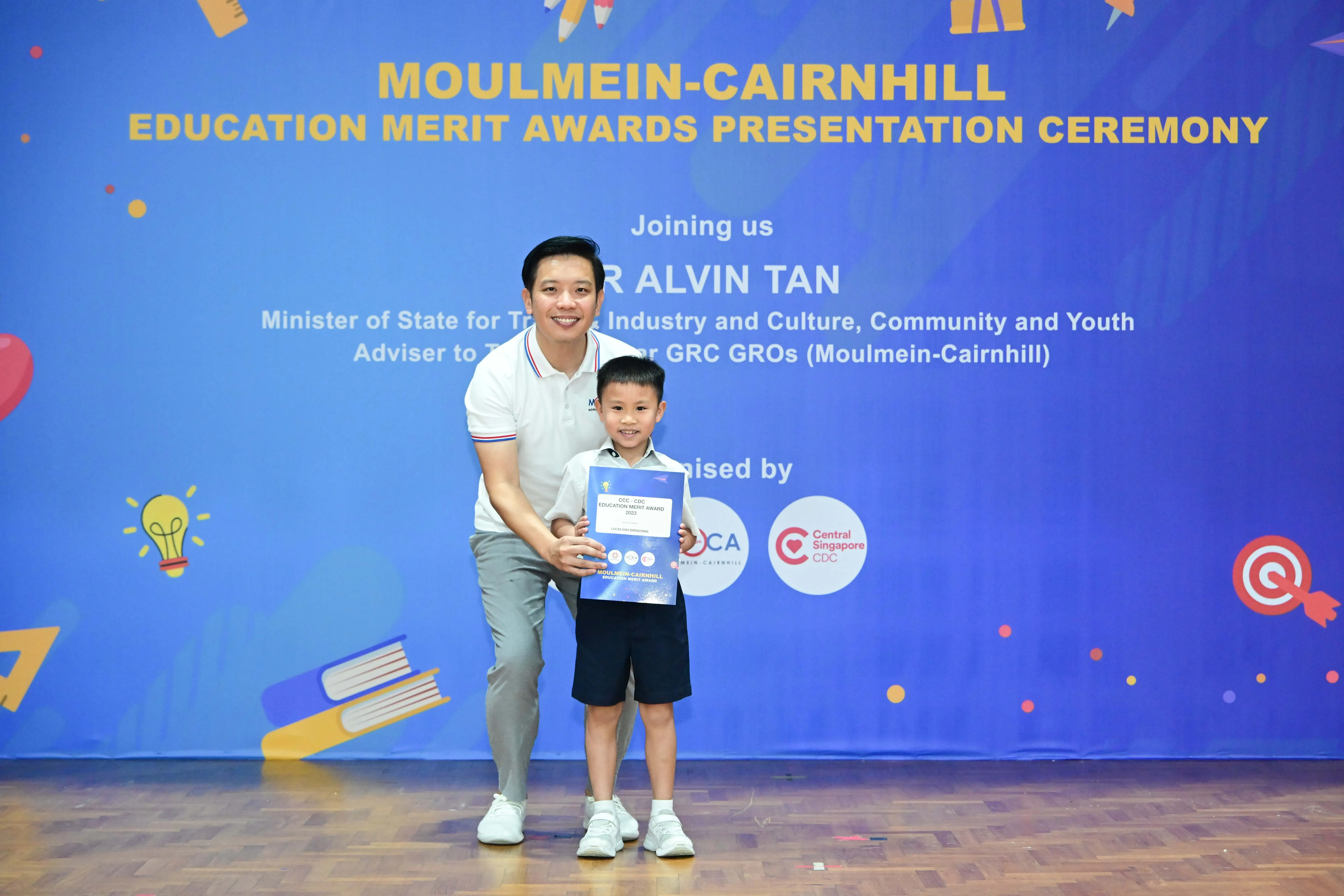 A man and young boy hold a certificate in front of a blue Moulmein-Cairnhill Education Merit Awards banner.