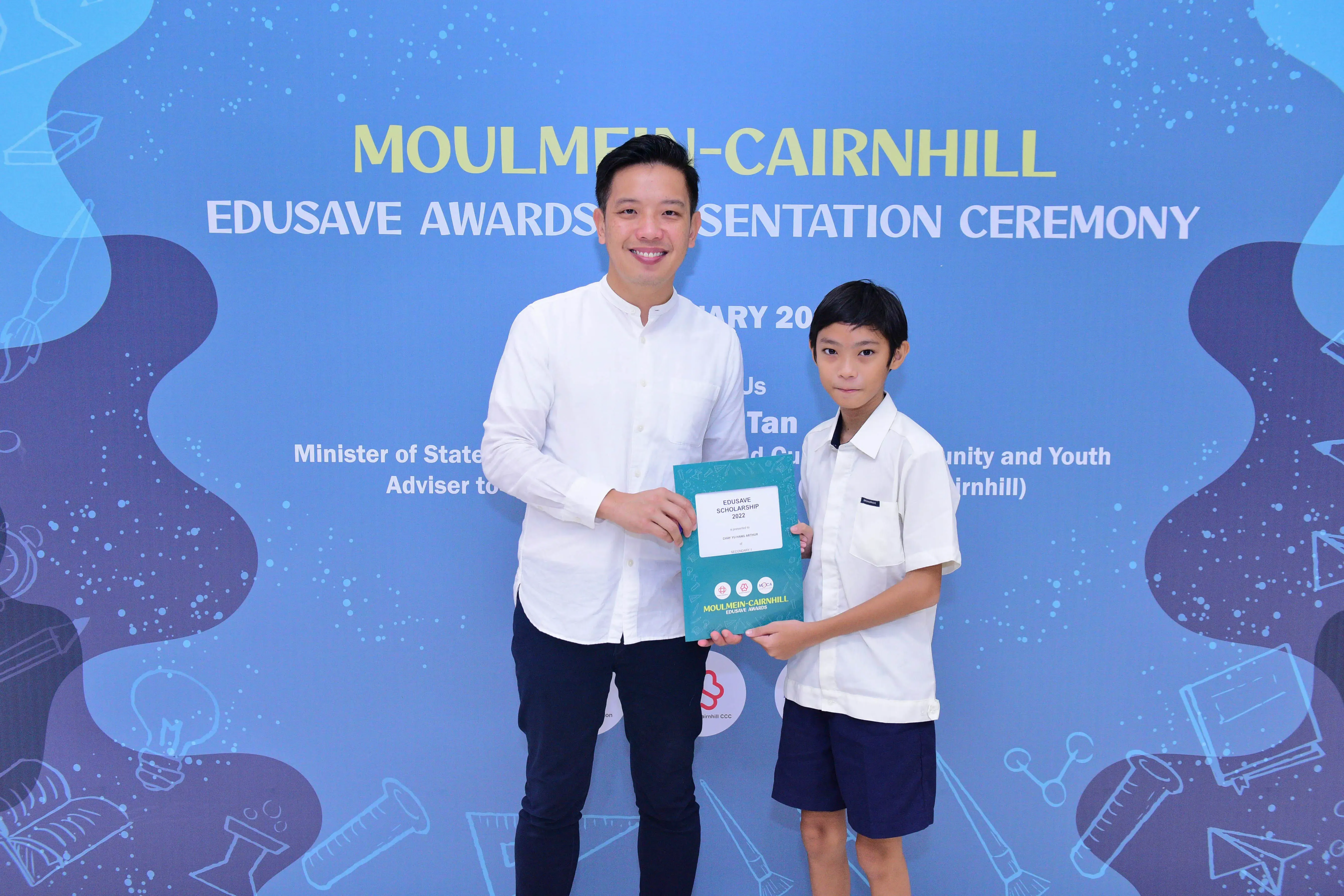 An adult man and a young boy hold a teal certificate together at the Moulmein-Cairnhill Edusave Awards Ceremony.