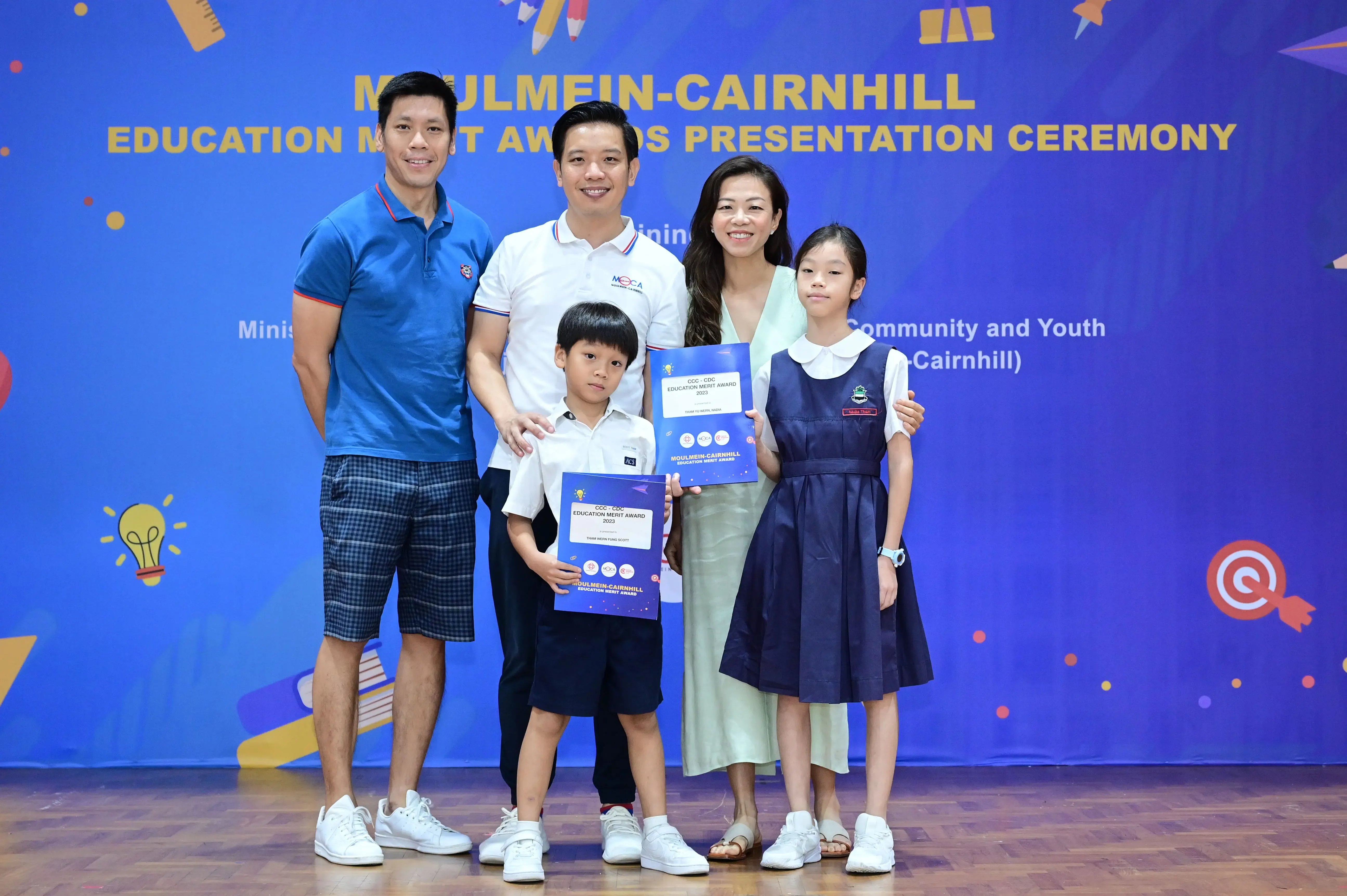 Five people pose in front of a Moulmein-Cairnhill Education Merit Awards banner, two children holding award certificates.