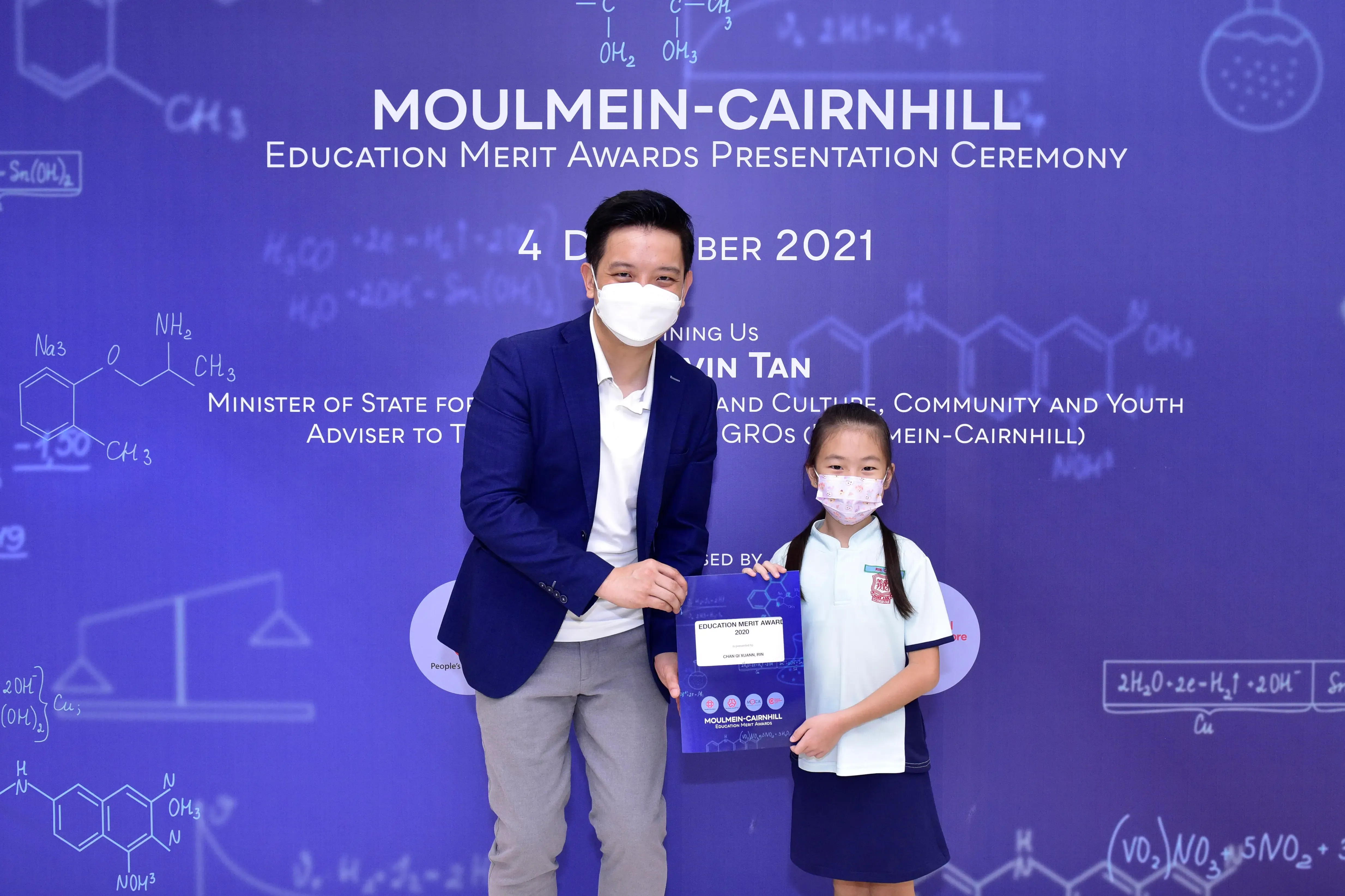 A masked man and young girl hold an Education Merit Award certificate at the Moulmein-Cairnhill ceremony, December 2021.