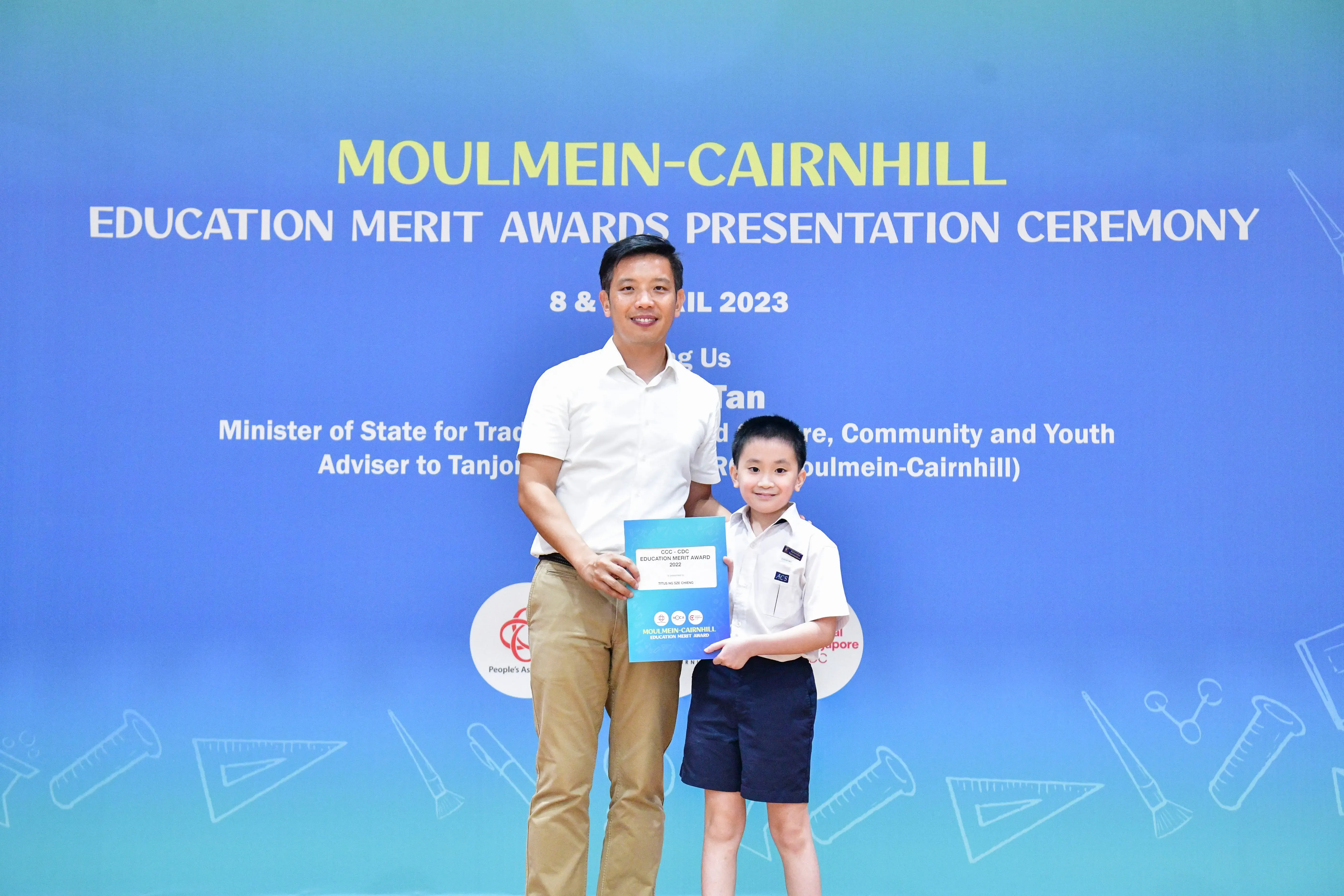 A man and a young boy holding a certificate stand before a Moulmein-Cairnhill Education Merit Awards 2023 banner.