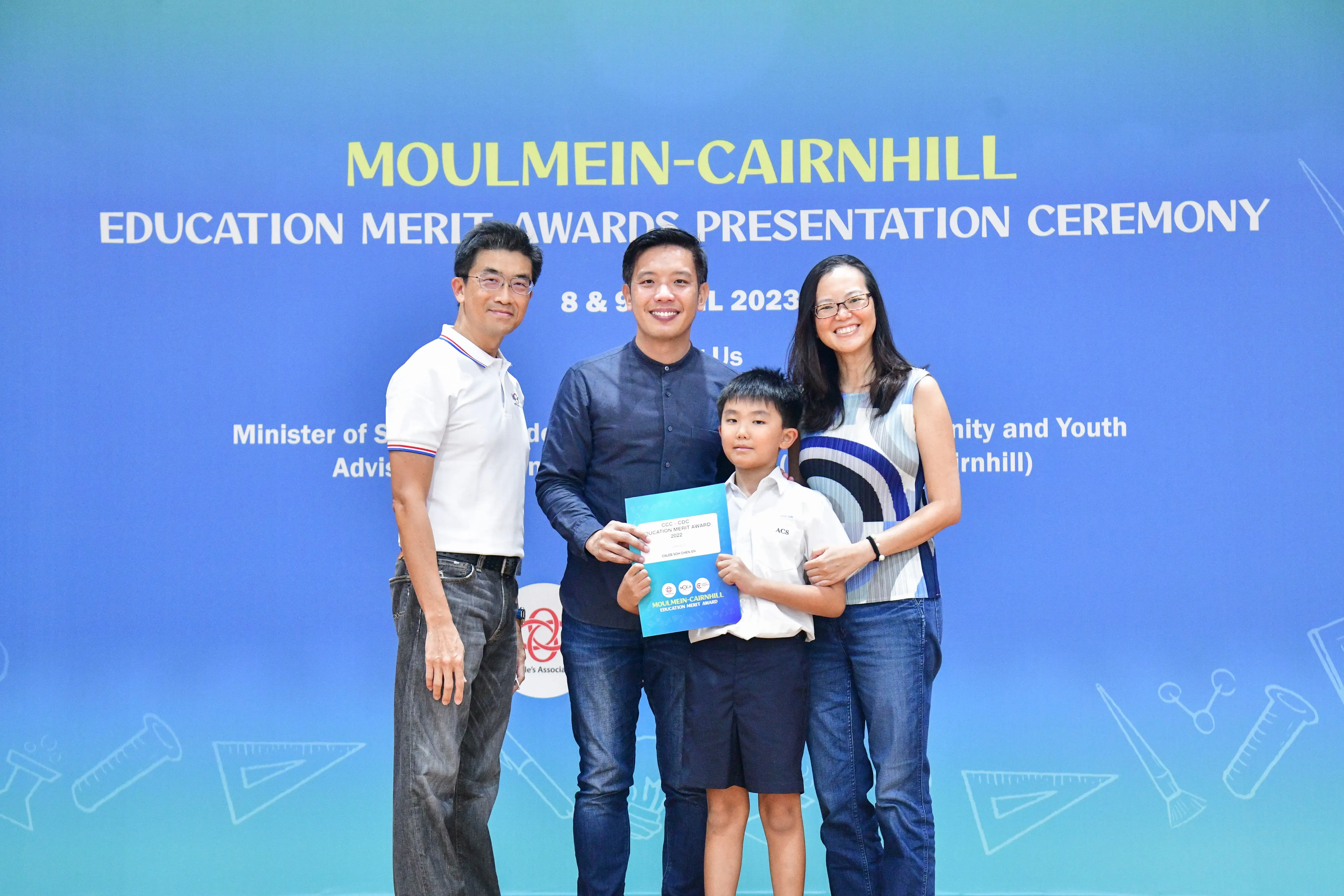 Four people pose before a Moulmein-Cairnhill Education Merit Awards banner; a boy holds a blue certificate.