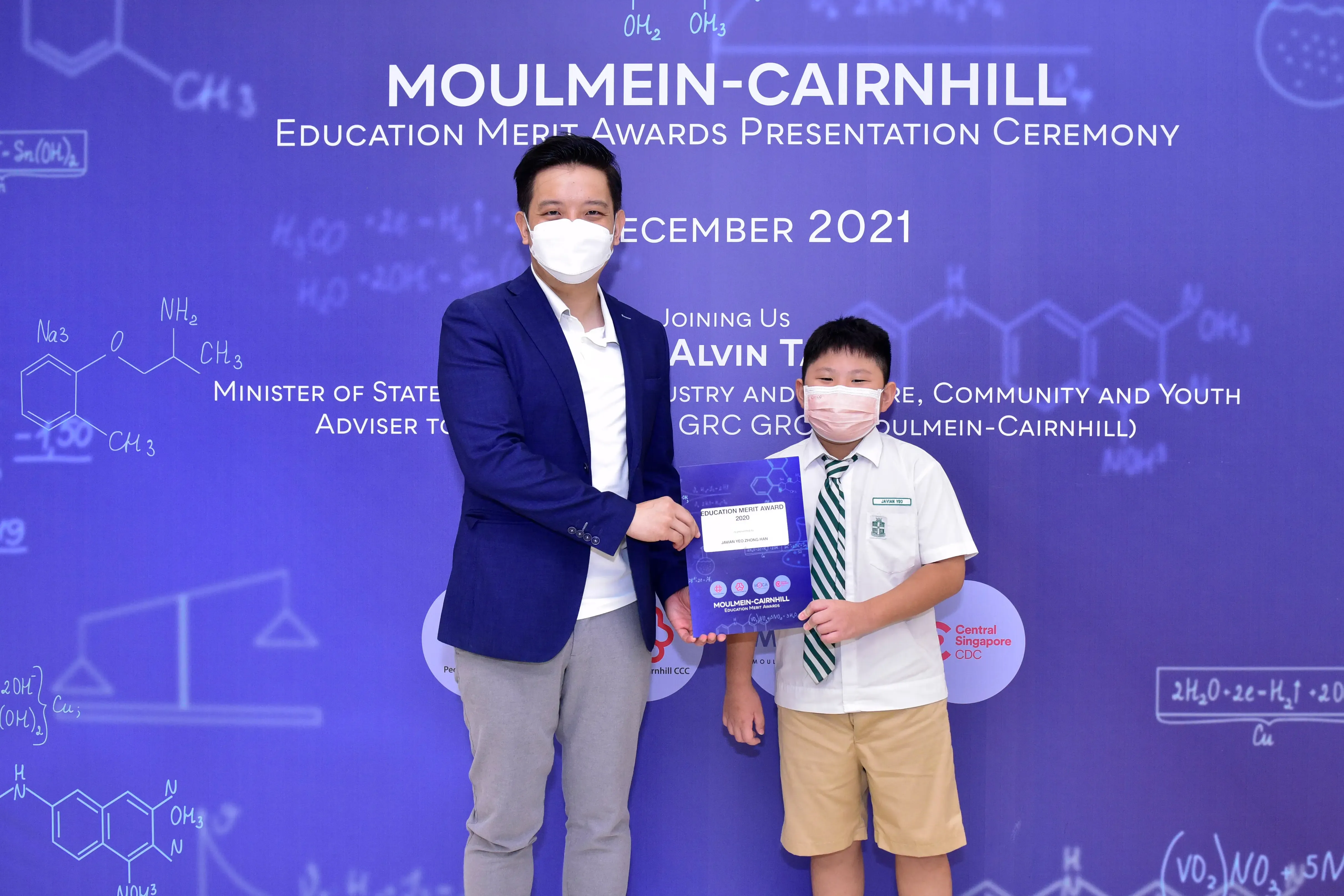 An adult in a blue blazer presents a certificate to a young boy in school uniform at a 2021 award ceremony.