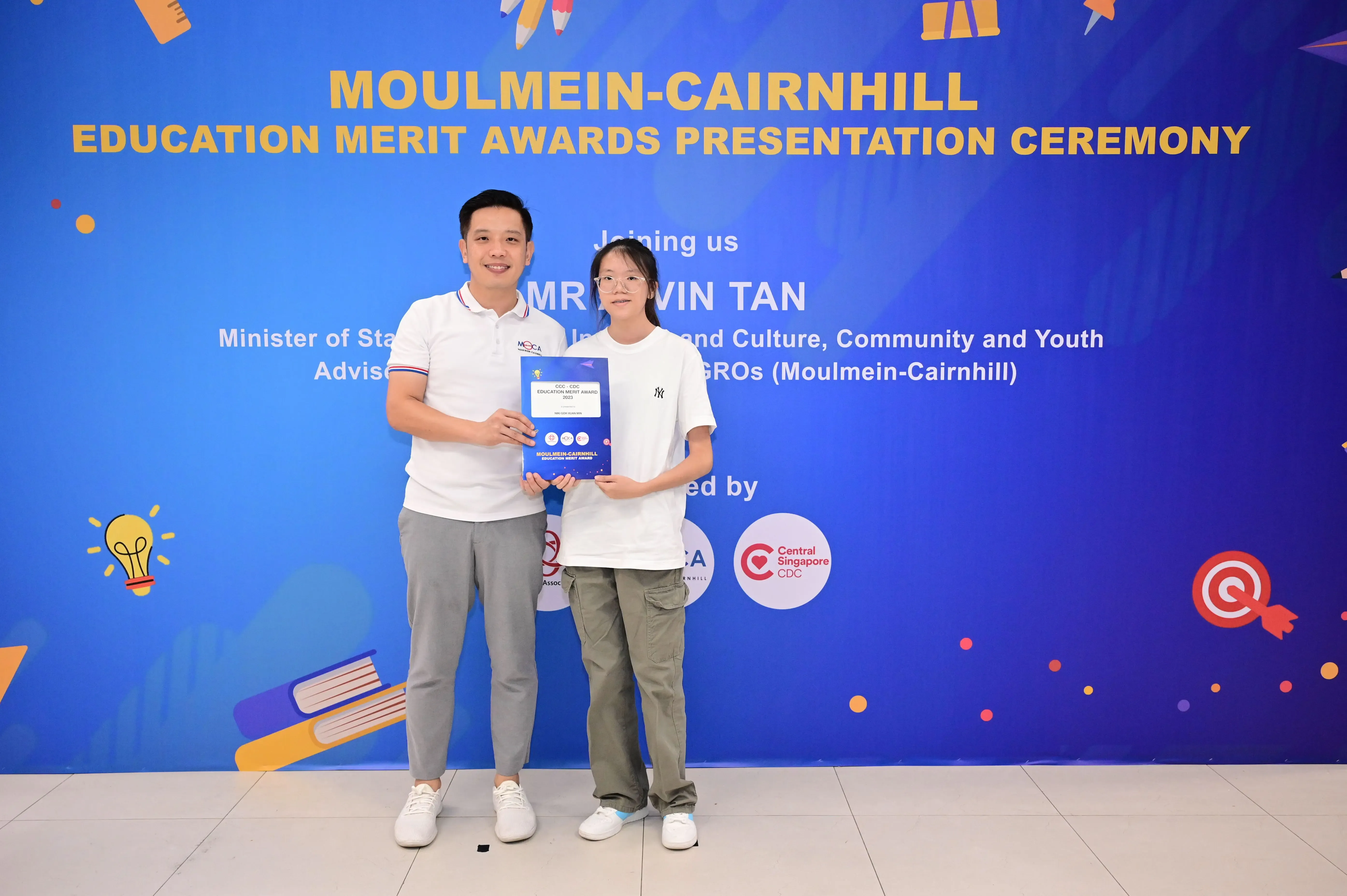 Two people hold an award booklet before a Moulmein-Cairnhill Education Merit Awards Presentation Ceremony banner.