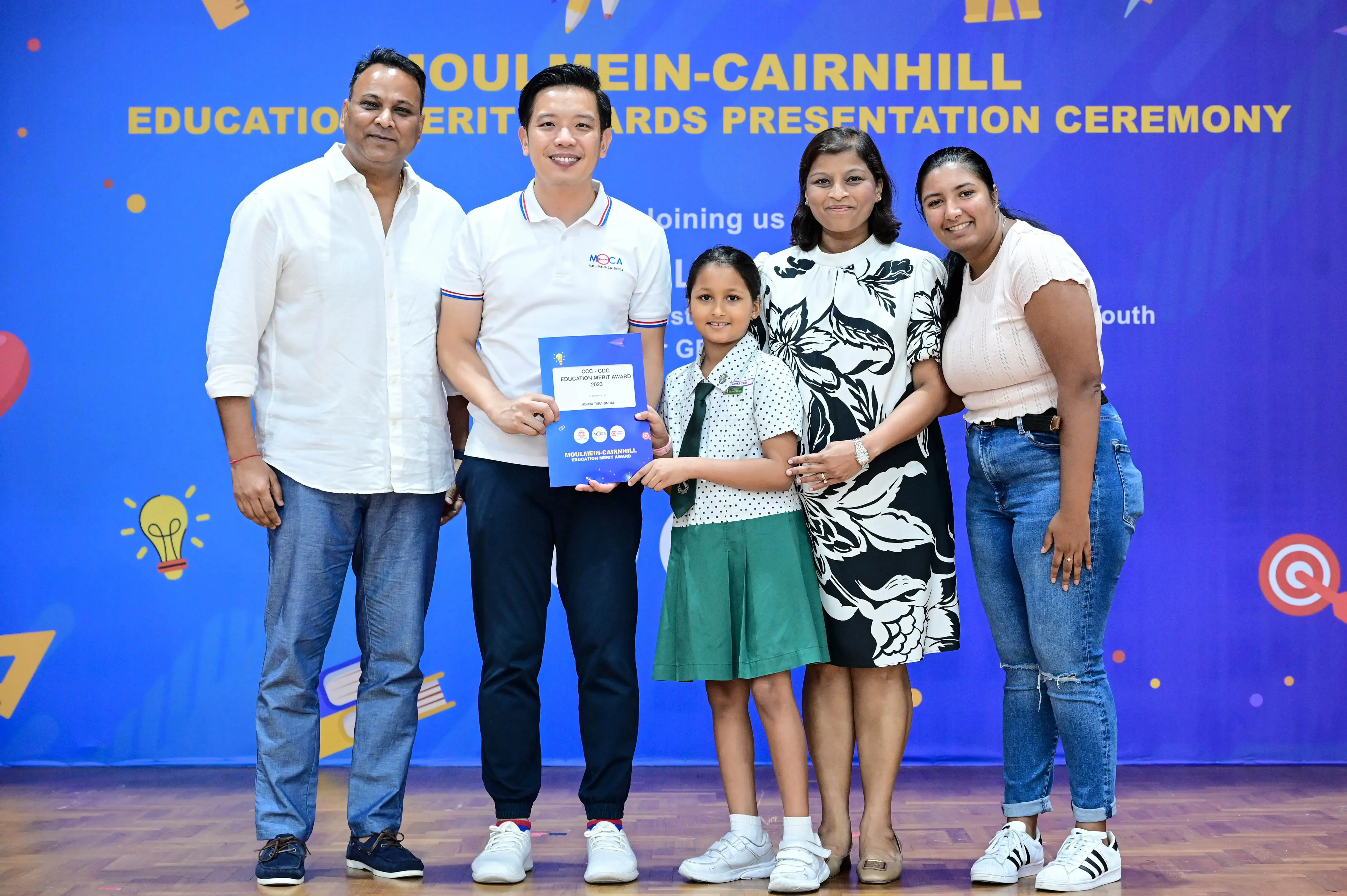 Four adults and a young girl in school uniform hold an award certificate at the Moulmein-Cairnhill Education Merit Awards ceremony.