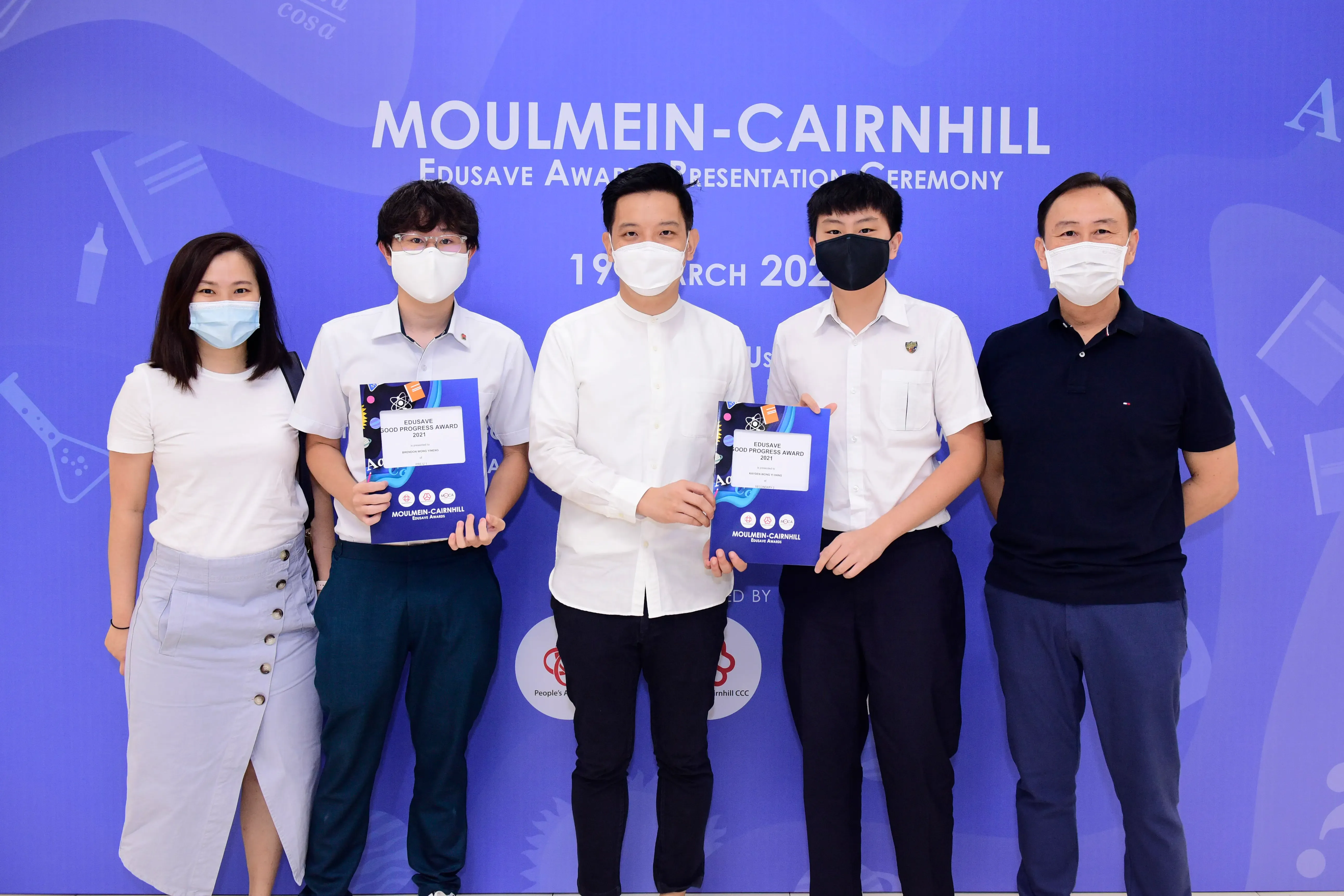 Five masked people pose at the Moulmein-Cairnhill Edusave Award Presentation Ceremony, two holding blue award certificates.